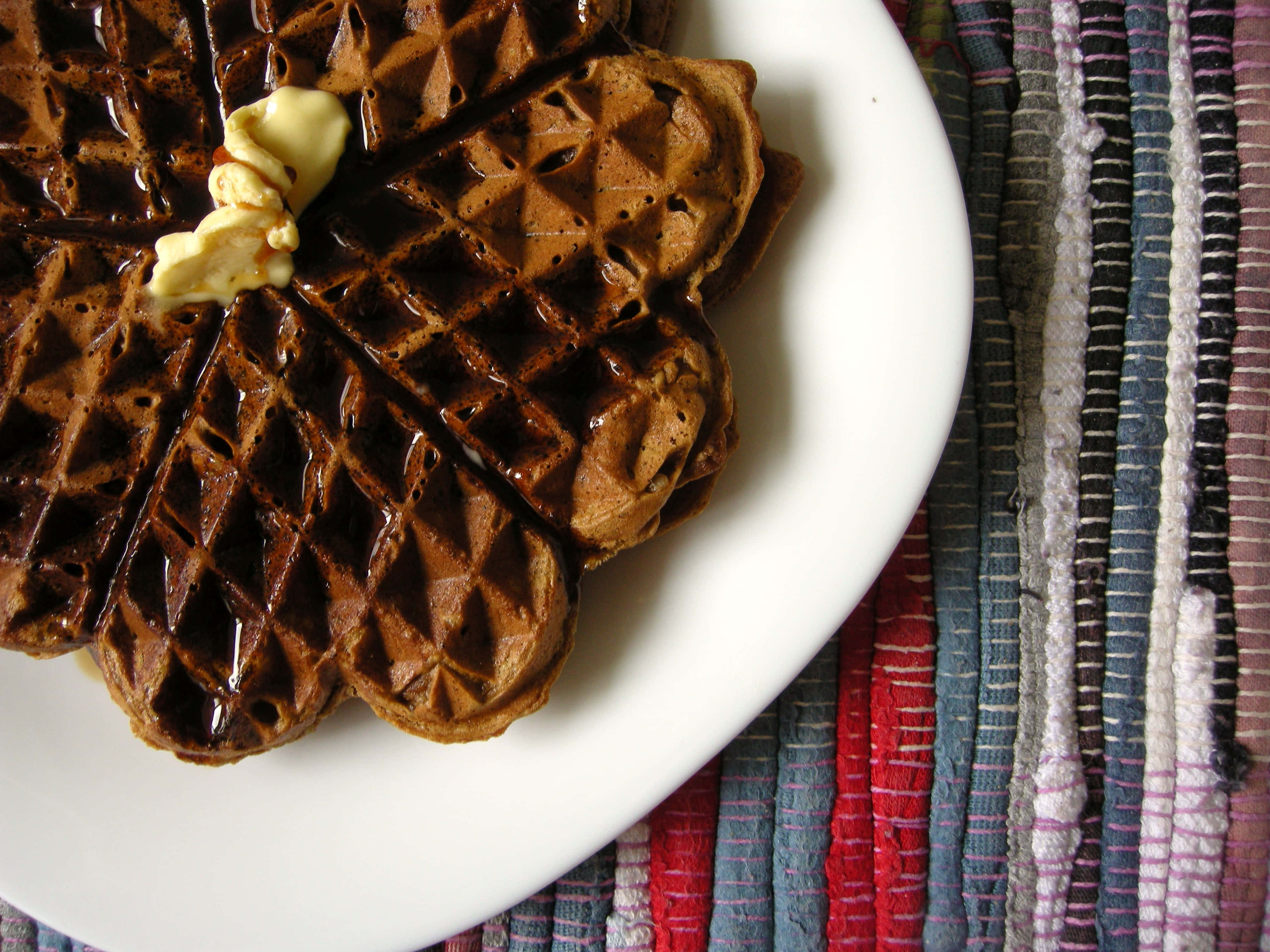 Waffle 桌布 壁紙 — 金黃色窩夫淋上糖漿，頂部放著融化的牛油，放在白色碟子上，背景是色彩繽紛的條紋紡織品。