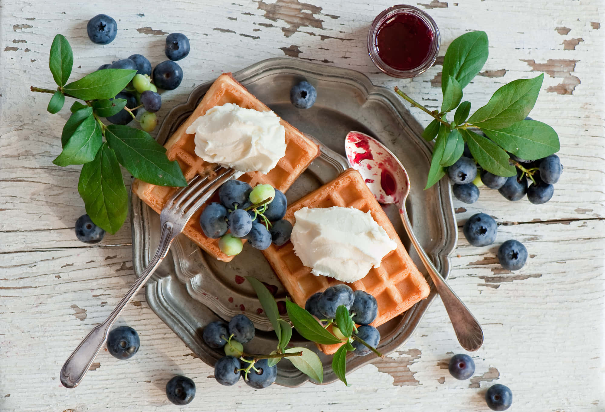 Waffle 桌布 壁紙 — 金黃色窩夫配上忌廉和新鮮藍莓，放在一個暗淡的銀托盤上，周圍有綠葉和一罐莓果醬。