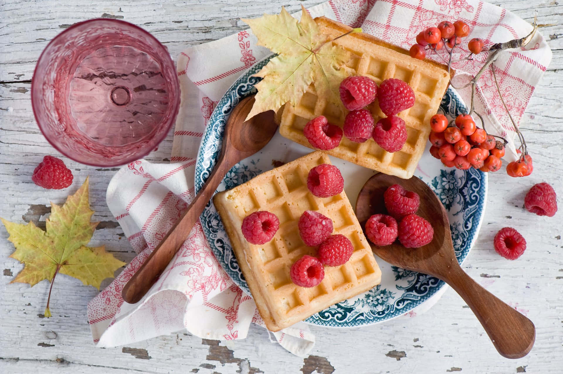 Waffle 桌布 壁紙 — 金黃色窩夫配上新鮮紅桑莓和秋葉，放在有圖案的碟上，旁邊有粉紅色玻璃杯和木匙。