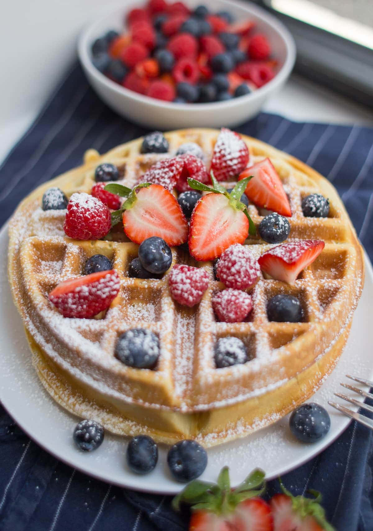 Waffle 桌布 壁紙 — 金黃色窩夫配上新鮮草莓、藍莓和糖粉，放在白色碟上，背景有一碗雜莓。