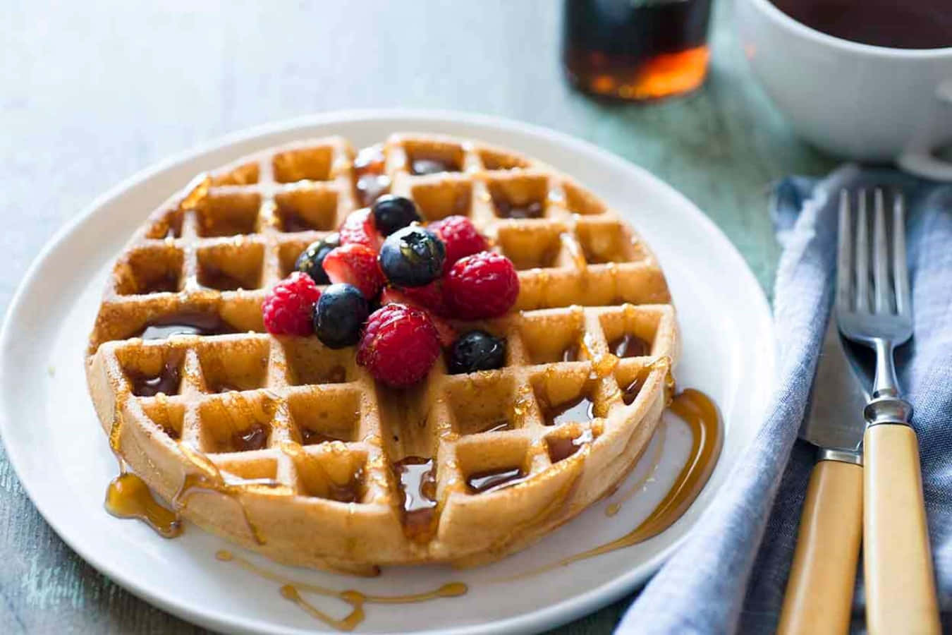 Waffle 桌布 壁紙 — 金黃色窩夫配上新鮮莓果和糖漿，放在白色碟子上，旁邊有杯子和餐具。