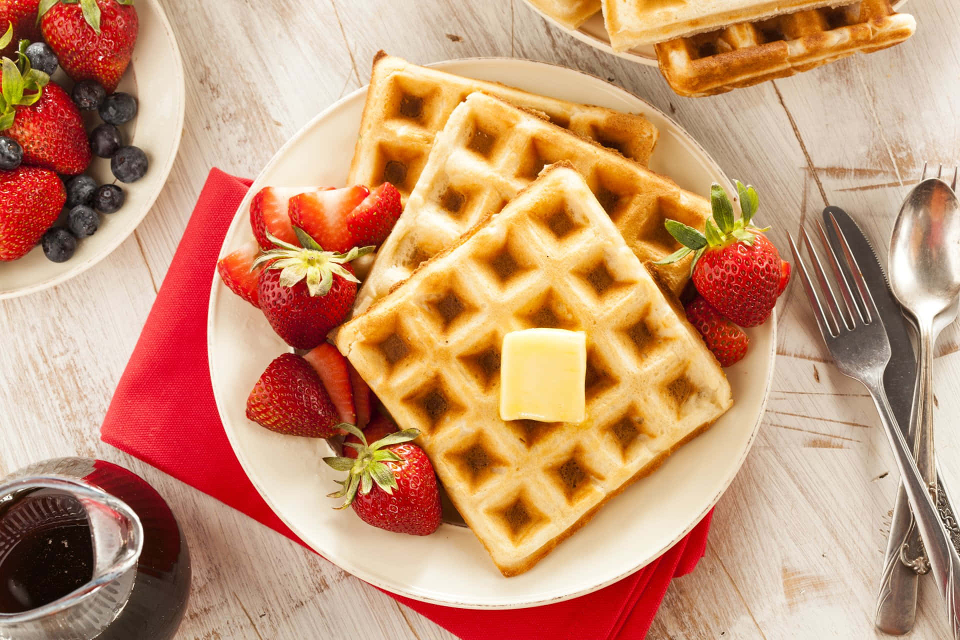 Waffle 桌布 壁紙 — 金黃色窩夫配上牛油和新鮮草莓，放在白色碟上，旁邊有一碗莓果和糖漿，置於鄉村風木桌上。