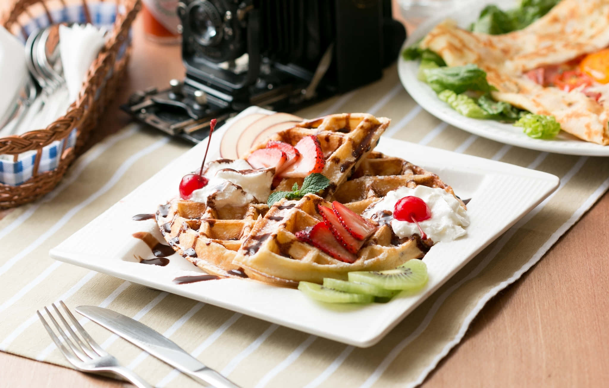 Waffle 桌布 壁紙 — 金黃色窩夫配上草莓、忌廉和奇異果片，放在白色碟上，旁邊有復古相機和可麗餅。