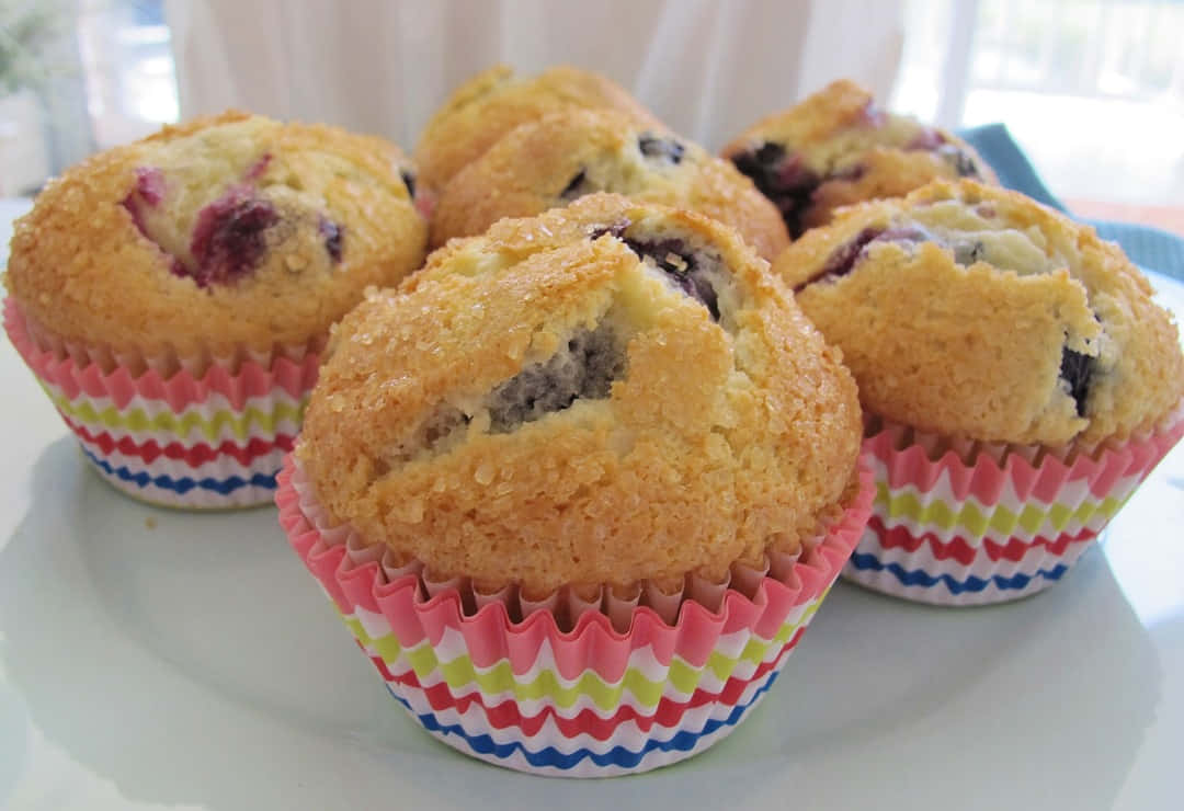 Blueberry Muffins 桌布 壁紙 — 金黃色藍莓鬆餅放在色彩繽紛的條紋紙托中，整齊地擺放在明亮的窗邊白色碟子上，令人垂涎。