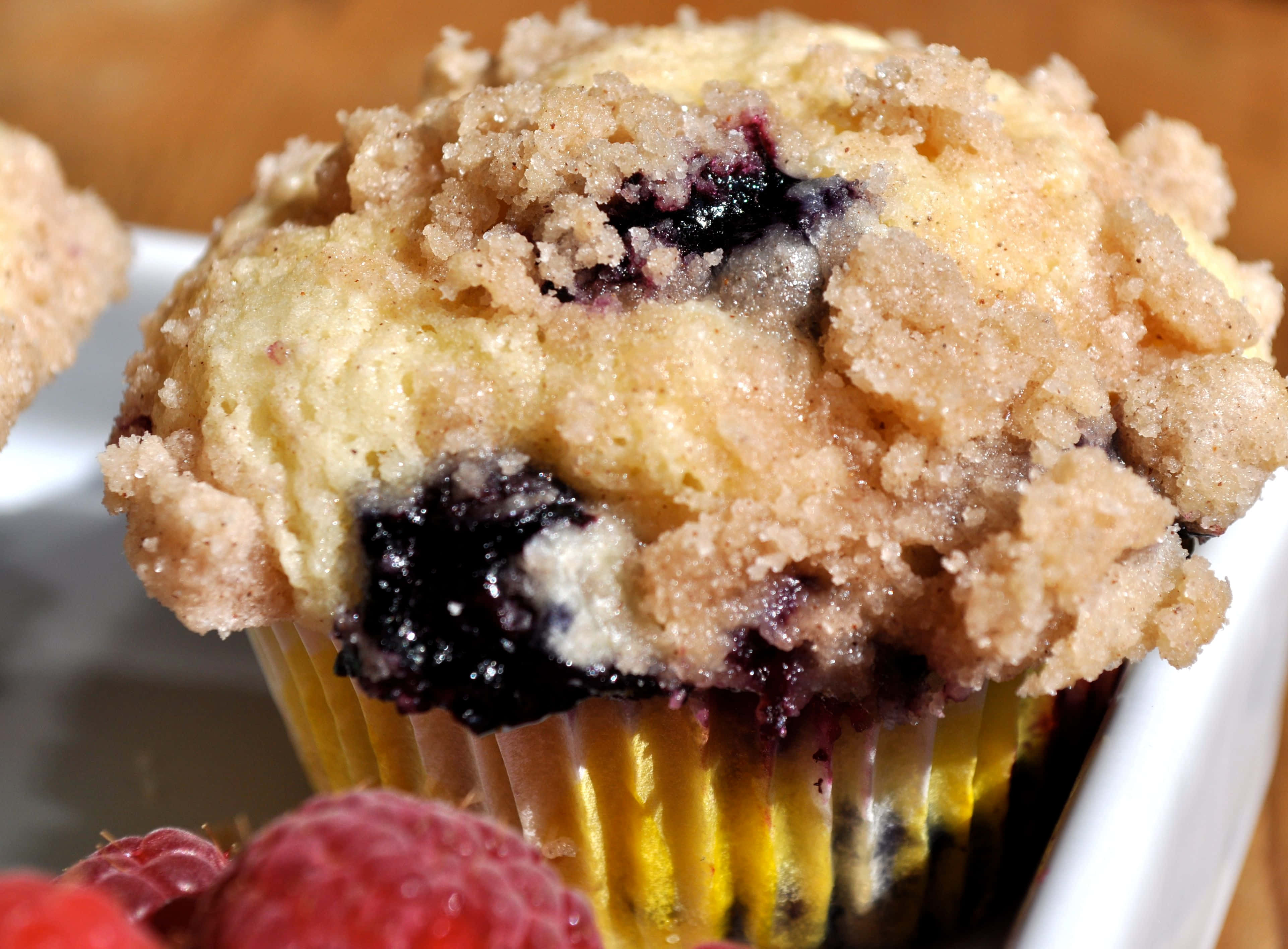 Blueberry Muffins 桌布 壁紙 — 金黃色藍莓鬆餅的近距離特寫，上面鋪滿酥脆的糖粒酥頂和新鮮覆盆子。