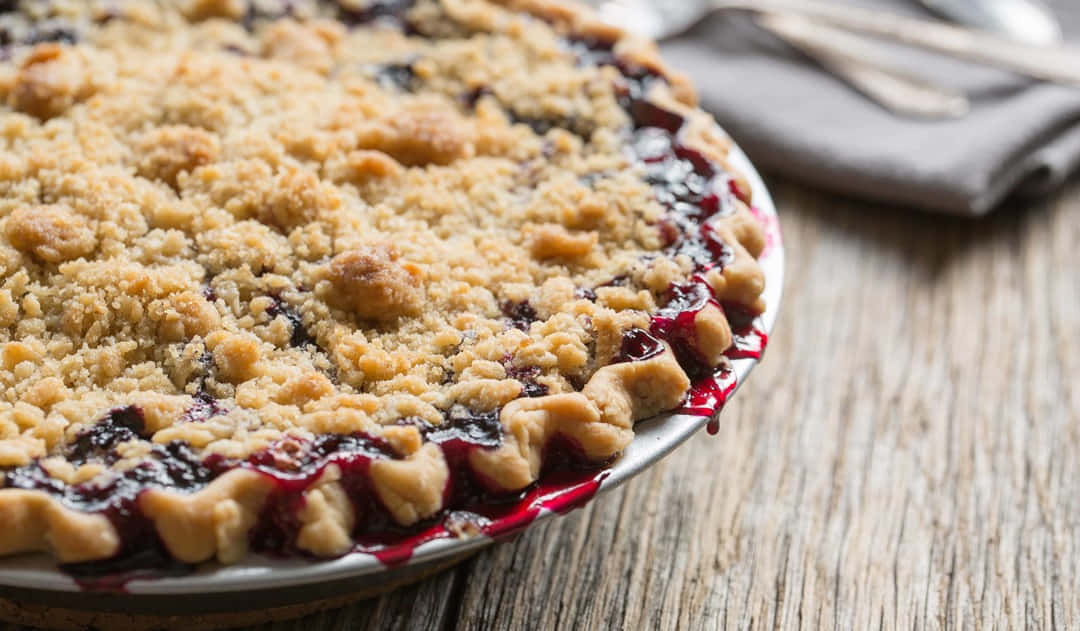 Blueberry Pie 桌布 壁紙 — 金黃色酥脆金寶藍莓批的特寫，果汁滲出在質樸的木桌上。