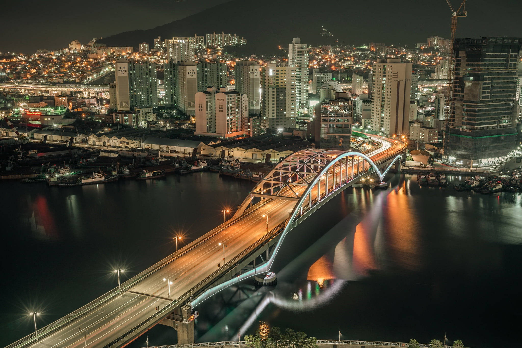 釜山大橋 壁紙 — 一座發光的拱橋在夜間橫跨黑暗的河流，在繁華的城市海濱反射出城市燈光。