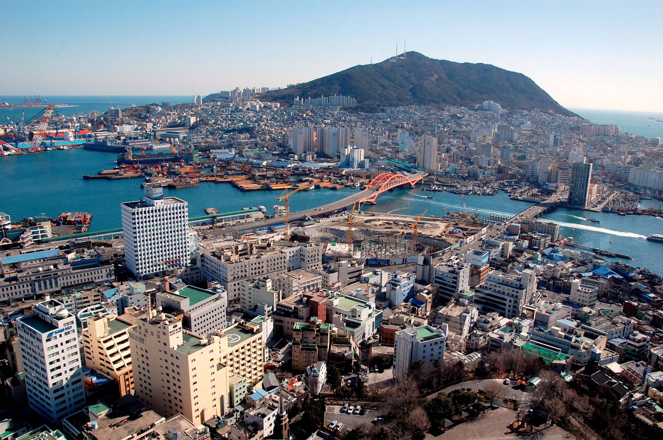 釜山水岸城市 壁紙 — 繁忙沿海城市的鳥瞰圖，一座紅色橋樑橫跨碧綠海水，密集的城市建築後方升起一座綠色山丘。