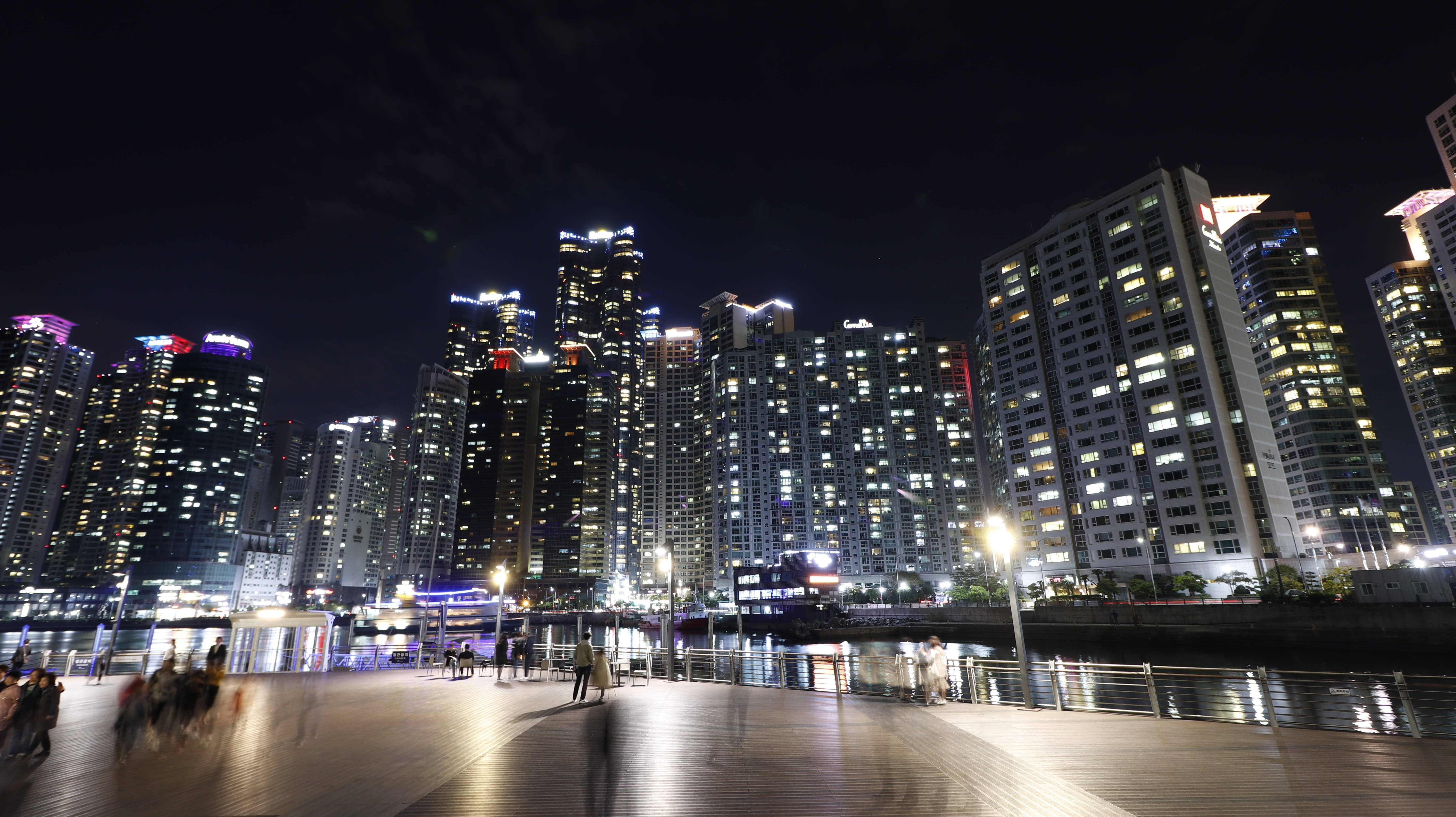 釜山馬里納城摩天大樓 壁紙 — 充滿活力的夜間城市景觀，燈火通明的摩天大樓倒映在水面上，木質長廊上模糊的行人。