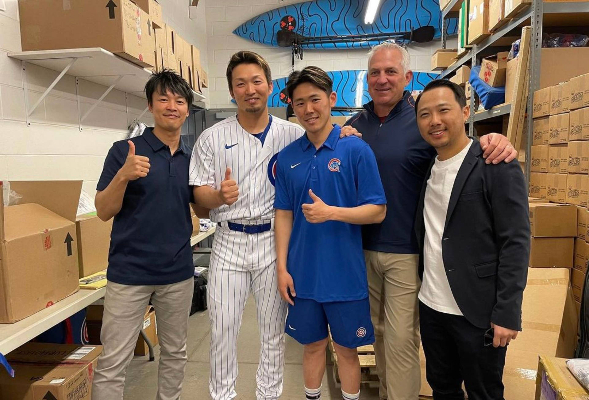 五個着住棒球制服同休閒服嘅男人喺倉庫入面開心咁擺甫士。