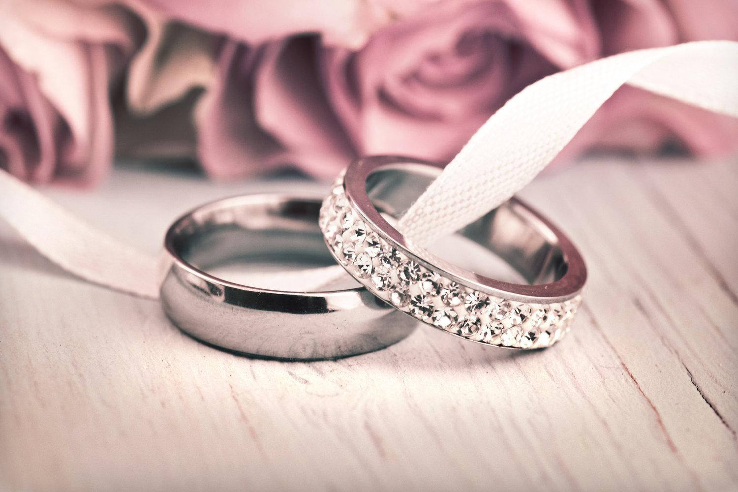 銀婚戒與白絲帶 壁紙 — 兩枚結婚戒指，一枚素面，一枚鑲鑽，放在白色絲帶上，旁邊是柔和的粉紅玫瑰，置於木質表面。