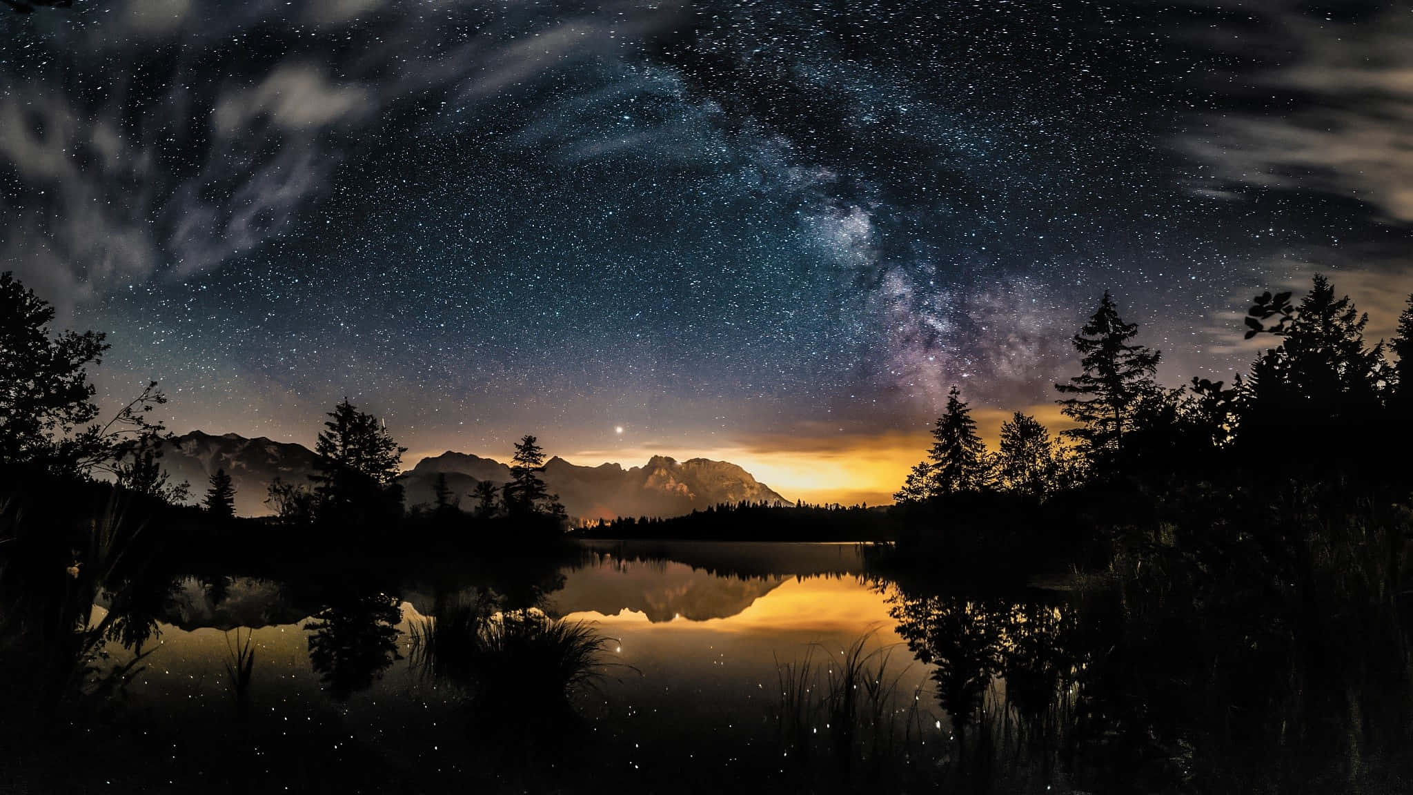 銀河橫跨湖面與樹林 壁紙 — 繁星點點的夜空，銀河拱起，映照在平靜湖面上的山脈和樹木剪影。