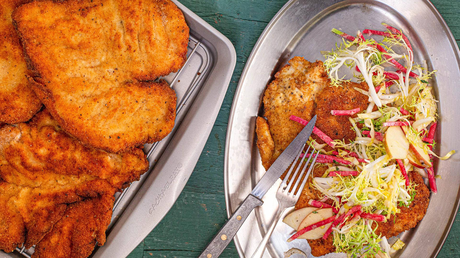 銀盤盛裝的維也納炸肉排 壁紙 — 金黃色炸肉排（Schnitzel）熱騰騰地放在托盤上，搭配色彩繽紛的苦苣沙律，置於鄉村風格的綠色木桌上。