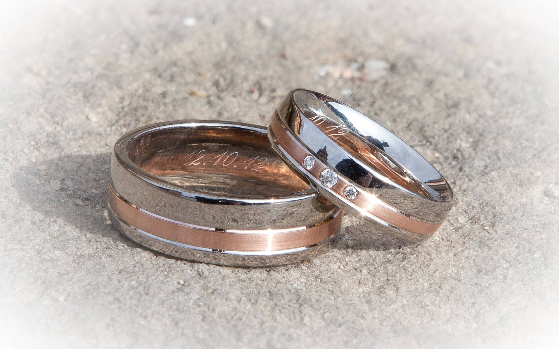 銀粉婚戒 壁紙 — 兩枚結婚戒指，其中一枚鑲有鑽石並刻有日期，放在紋理混凝土上，在明亮的陽光下。