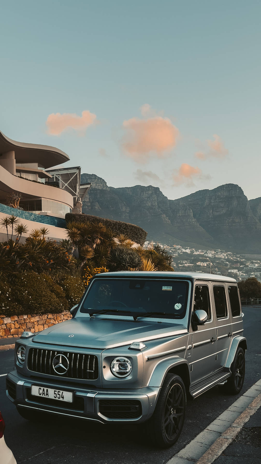 銀色 Mercedes G-Wagen 停泊在現代建築附近，背景是山脈和日落。