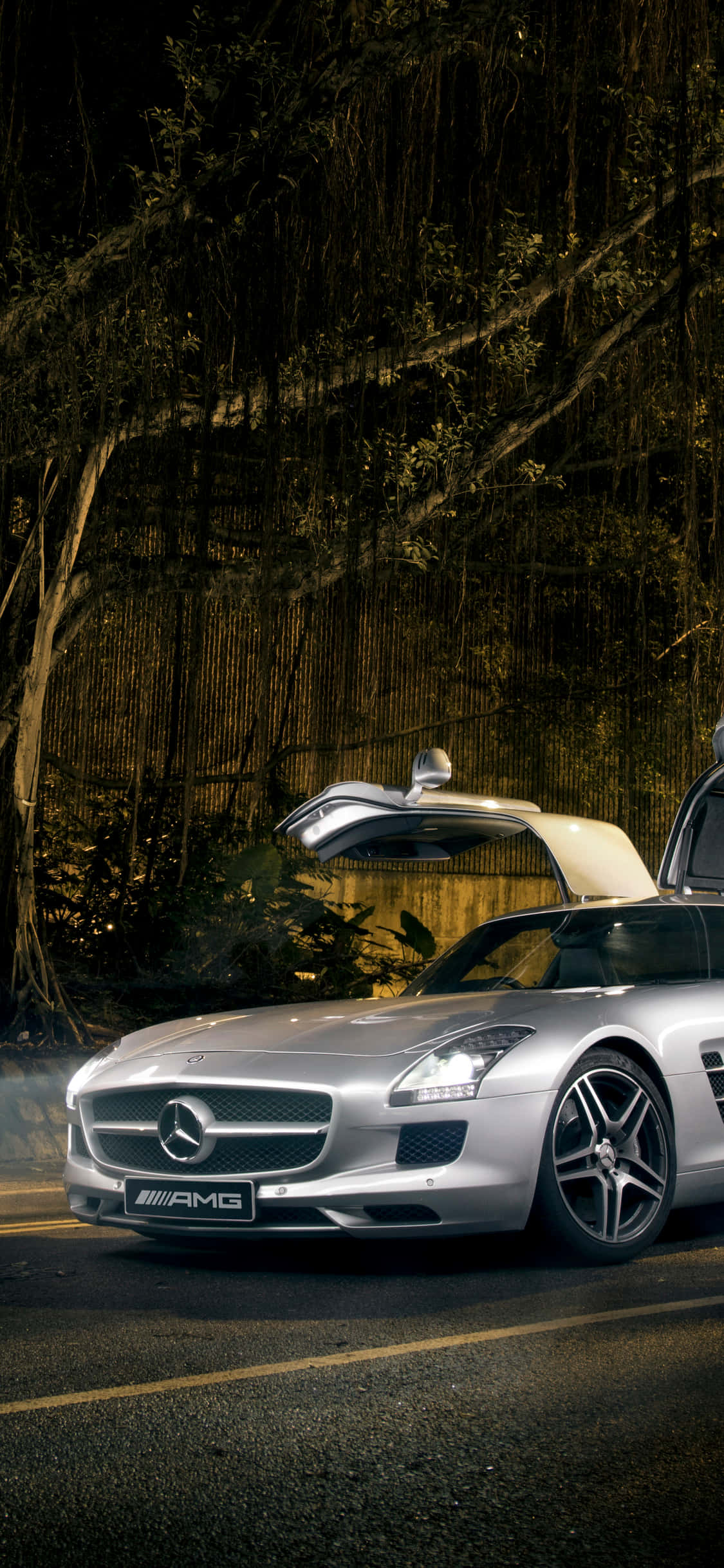 銀色 iPhone XS Mercedes AMG 背景 壁紙 — 一輛銀色平治AMG SLS跑車，鷗翼車門打開，在夜燈下閃耀，旁邊是長滿苔蘚的樹林道路。