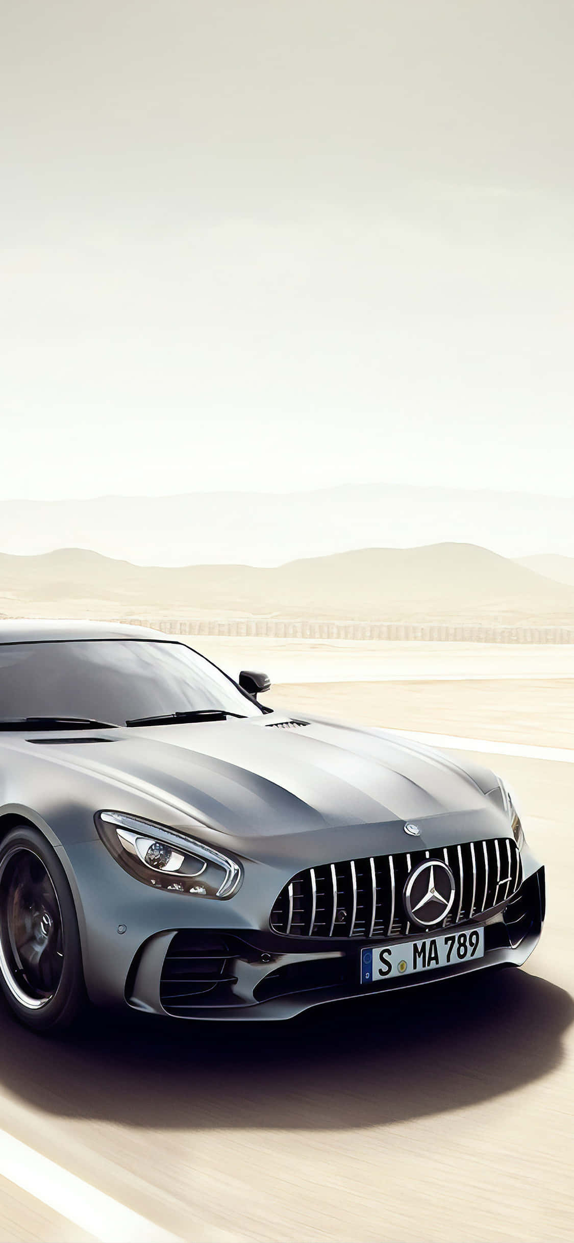 銀色 iPhone XS Mercedes AMG 沙漠背景 壁紙 — 一架流線型銀色 Mercedes-AMG GT 跑車在沙漠公路上飛馳，遠山在朦朧天空下若隱若現。