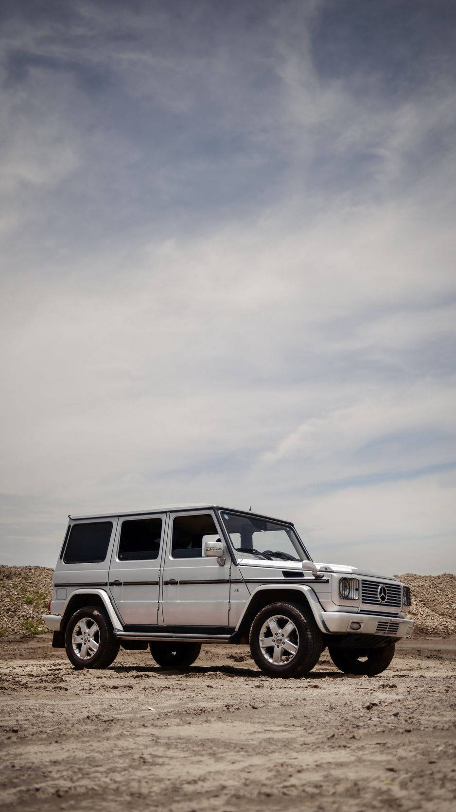 銀色 Mercedes-Benz G-Class SUV 停喺乾燥嘅泥地上，上面係多雲嘅天空。