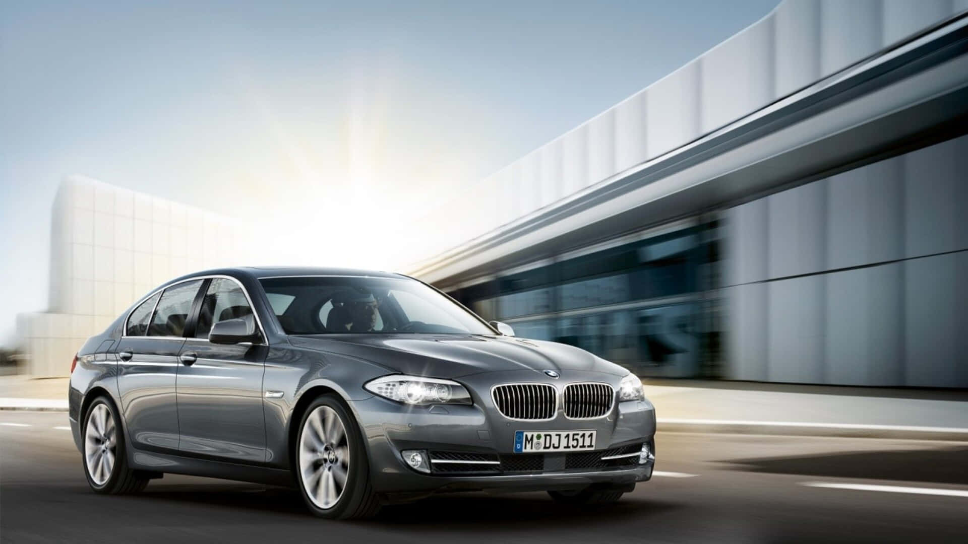 銀色BMW F10 1920x1080 M系列背景 壁紙 — 一輛時尚的銀色寶馬房車在明亮的日光下駛過現代玻璃建築。