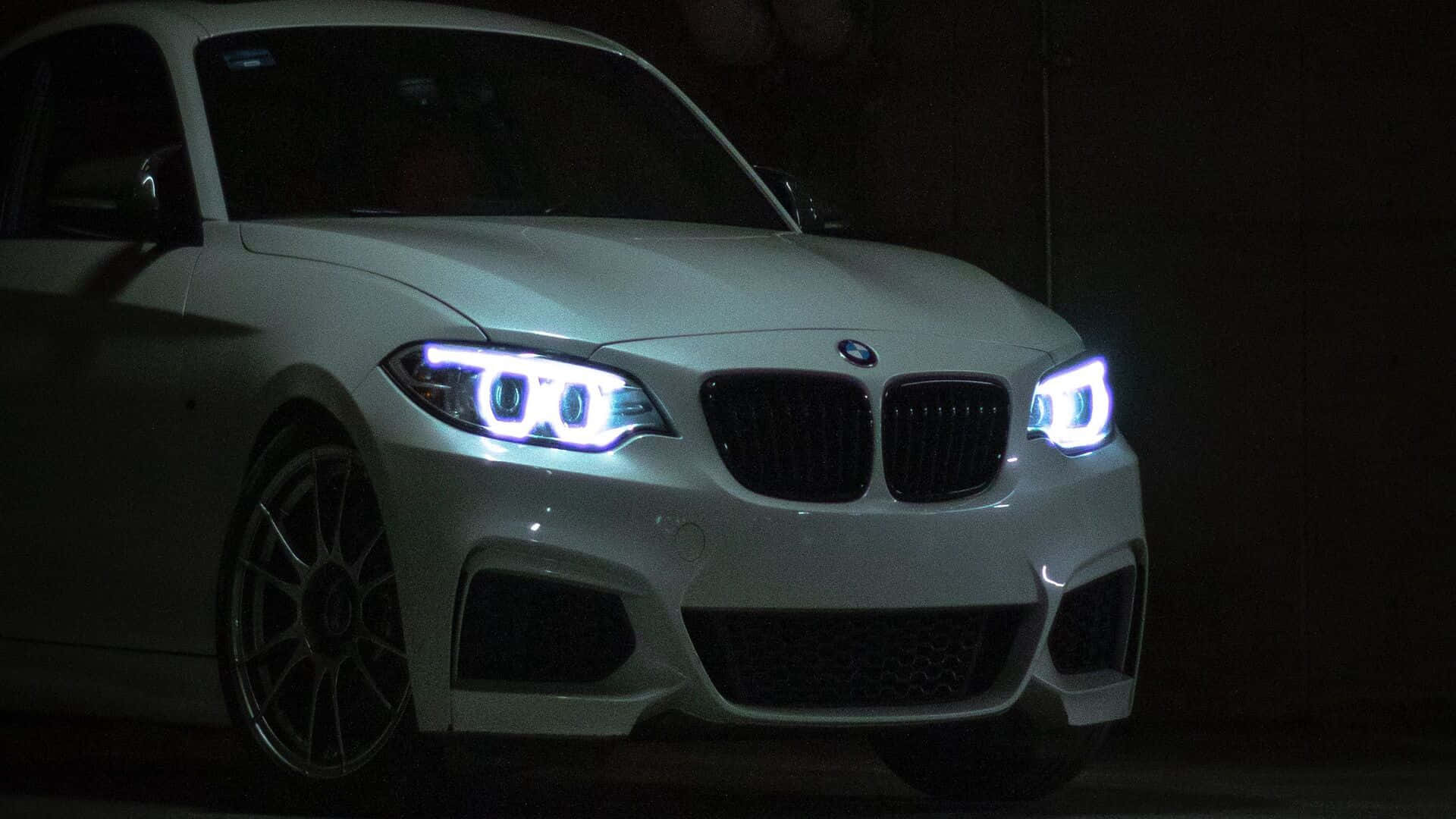 銀色BMW M2 1920x1080 M系列背景 壁紙 — 一輛白色BMW跑車在黑暗的車庫中發出明亮車頭燈的光芒，在其光滑表面投射出銳利的反射。