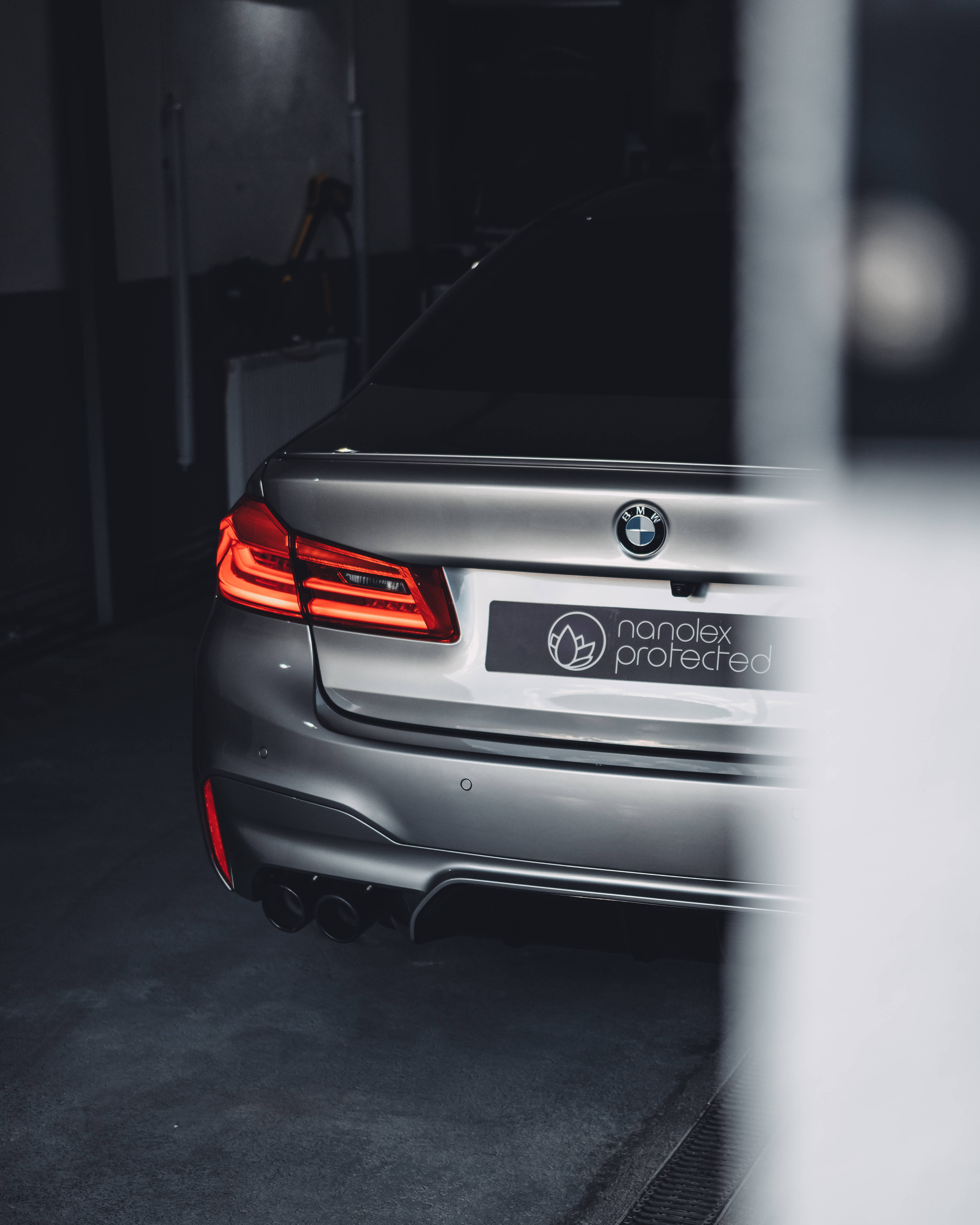 銀色BMW M車尾特寫 壁紙 — 一架時尚的銀色寶馬房車尾部在昏暗的車庫中發出紅色尾燈的光芒，並由模糊的前景元素框住。