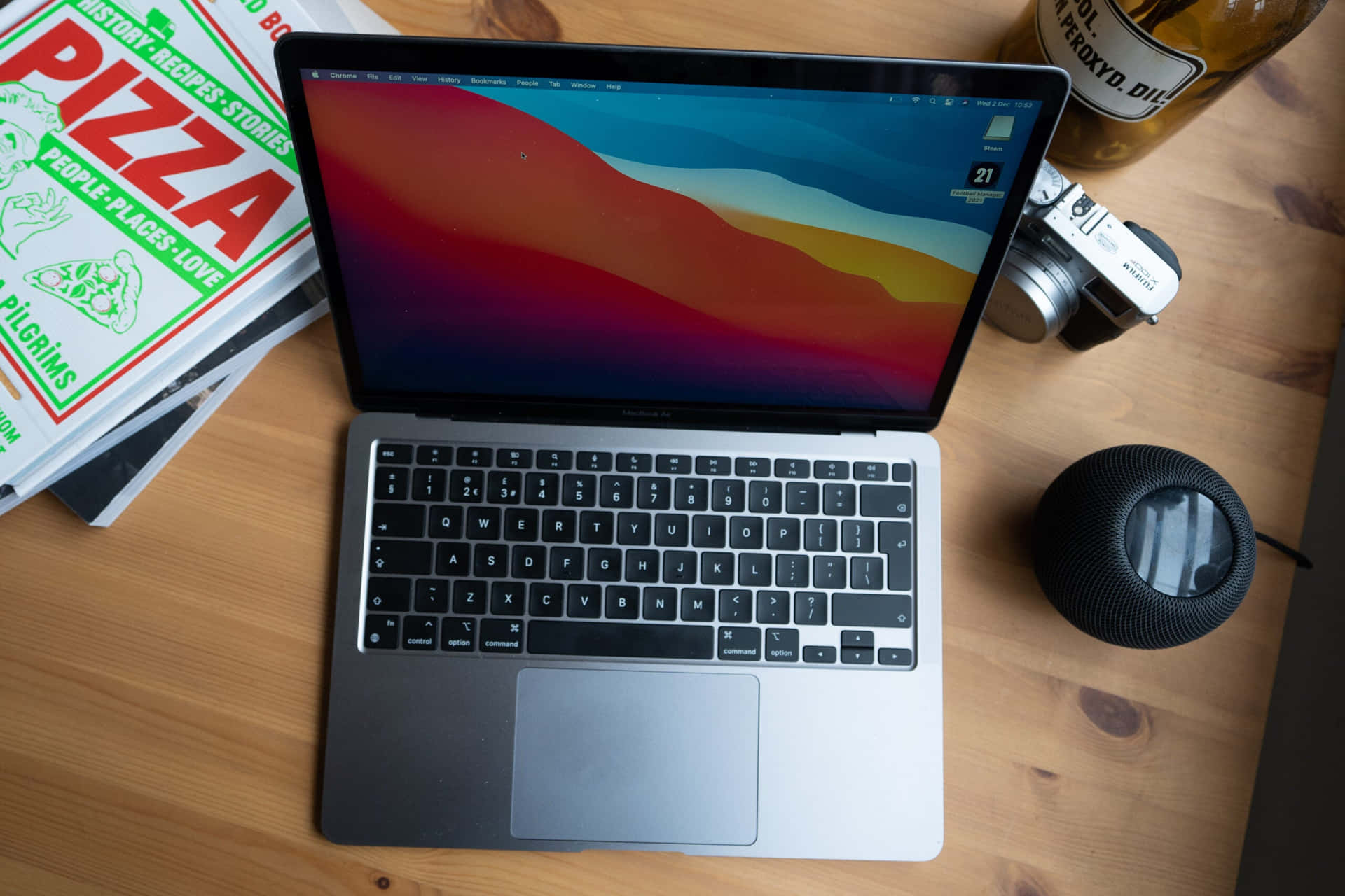 銀色MacBook搭配背光鍵盤 壁紙 — 一部銀色手提電腦帶有彩色抽象牆紙，放在木桌上，旁邊有相機、揚聲器和薄餅書。