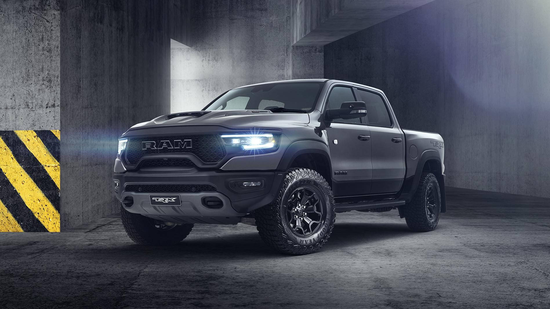 銀色RAM Truck 1500 TRX 壁紙 — 一輛啞光灰色的Ram TRX皮卡車，車頭燈發光，停在一個昏暗的混凝土車庫中，牆壁有黃黑條紋。