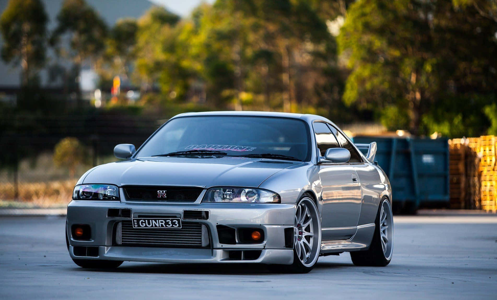 銀色低調R33 GTR 壁紙 — 一架銀色日產Skyline R34跑車，配備自訂輪圈及前保險桿，停泊在一個被樹木和垃圾箱包圍的混凝土場地上。