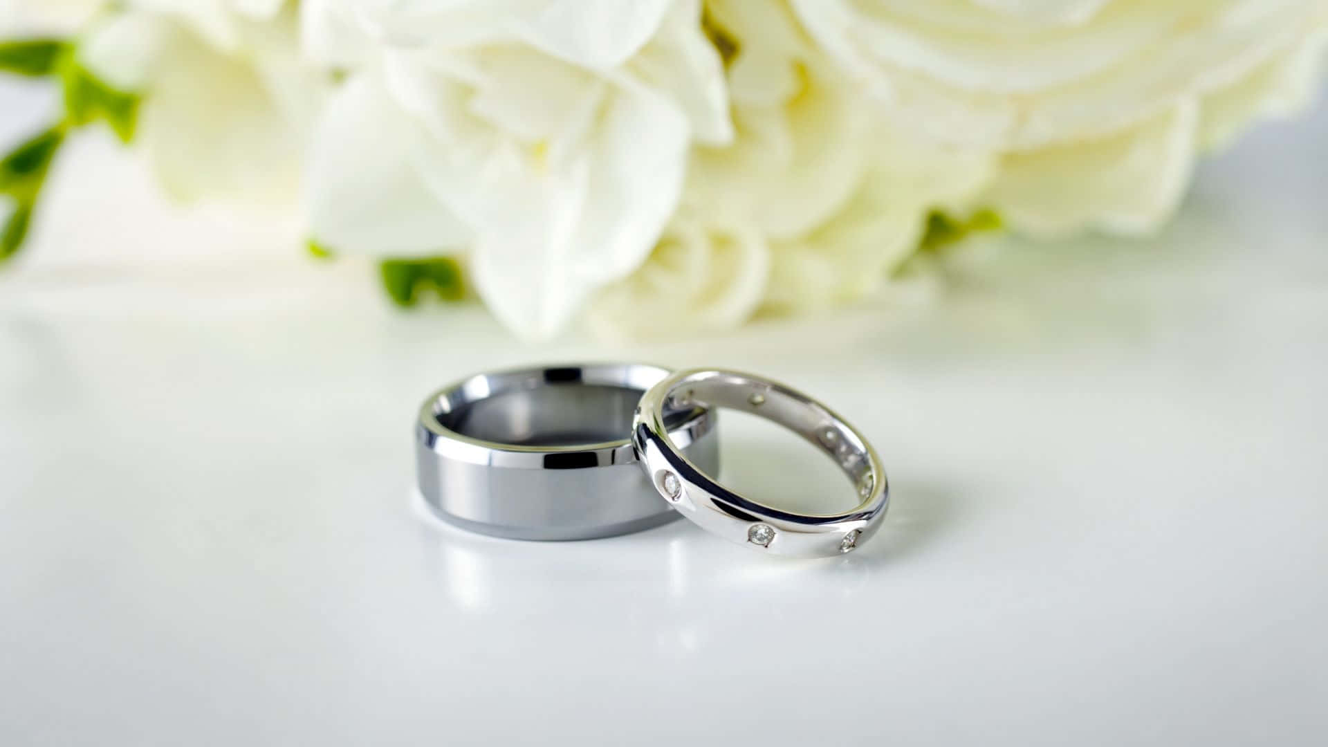 銀色婚戒配白花 壁紙 — 兩枚時尚的銀色結婚戒指放在白色表面上，旁邊是一束柔焦的白玫瑰。