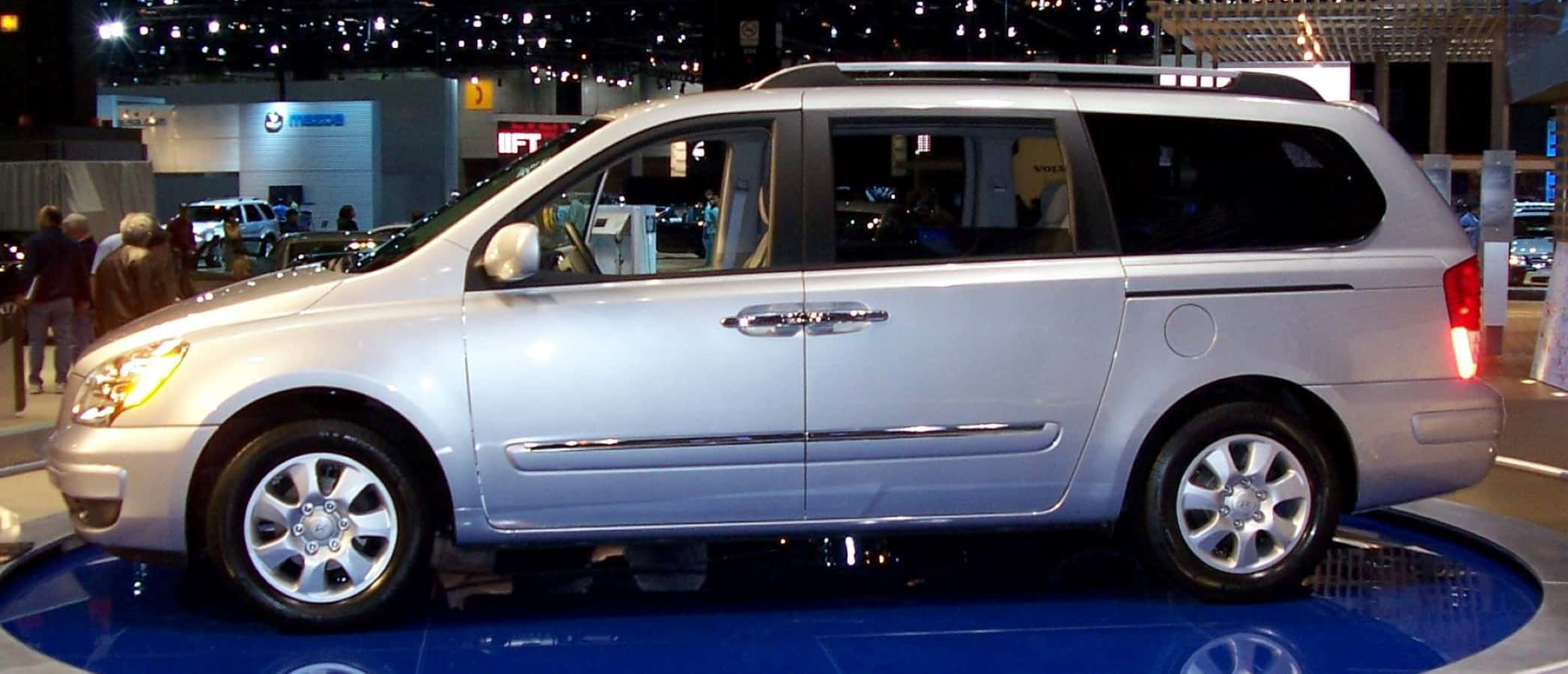 Hyundai Entourage 桌布 壁紙 — 銀色小型貨車在藍色旋轉平台上展示，明亮的展覽燈光下，背景有模糊的與會者。