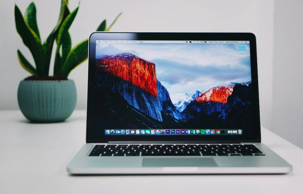 Laptop 桌布 壁紙 — 銀色手提電腦在白色桌子上，旁邊有一盆綠色植物，顯示著熾熱的日落山脈壁紙。
