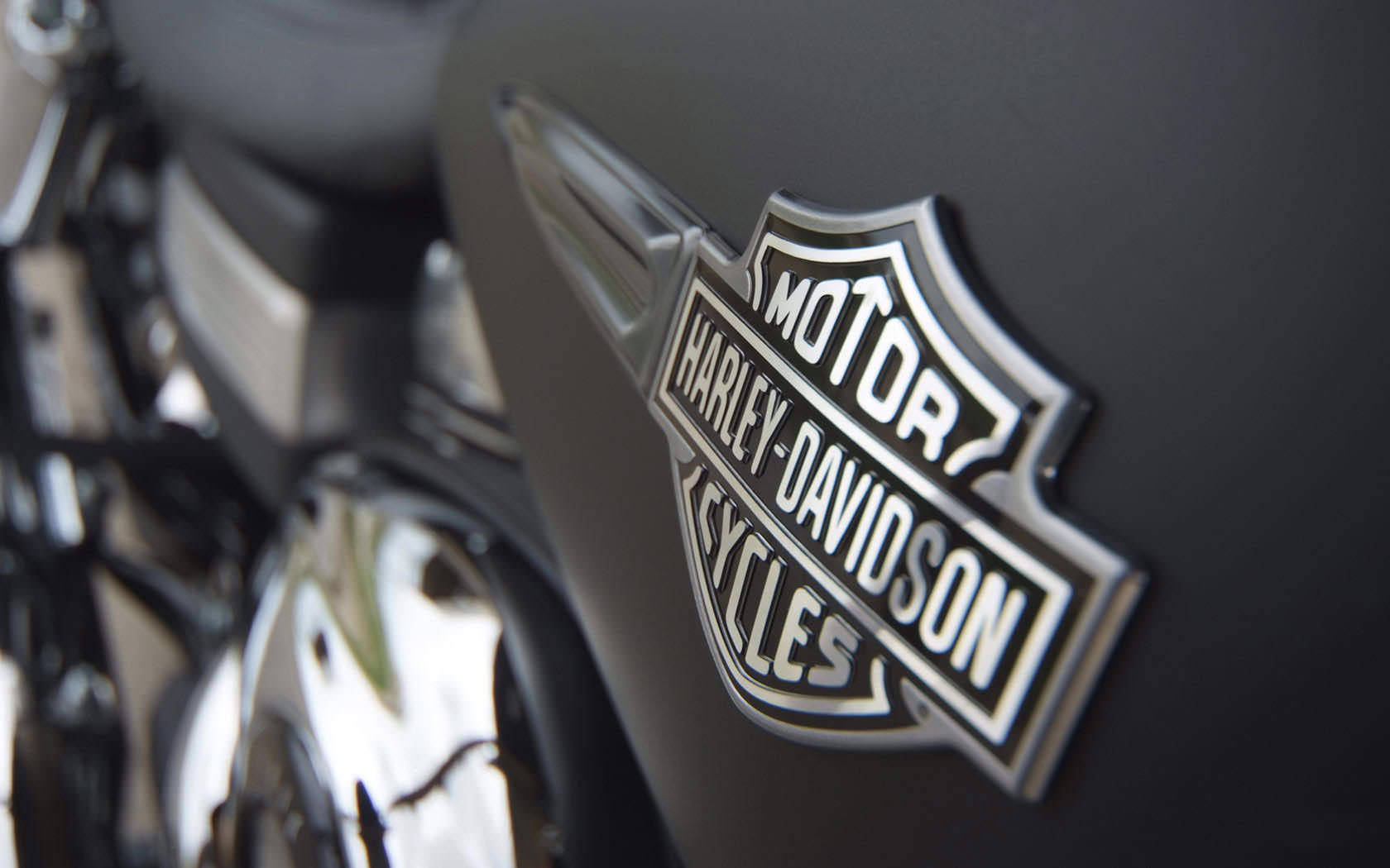 銀色板面哈雷戴維森標誌 壁紙 — 啞光黑色電單車油缸上嘅鉻 Harley-Davidson 標誌特寫，背景有模糊嘅鉻引擎零件。