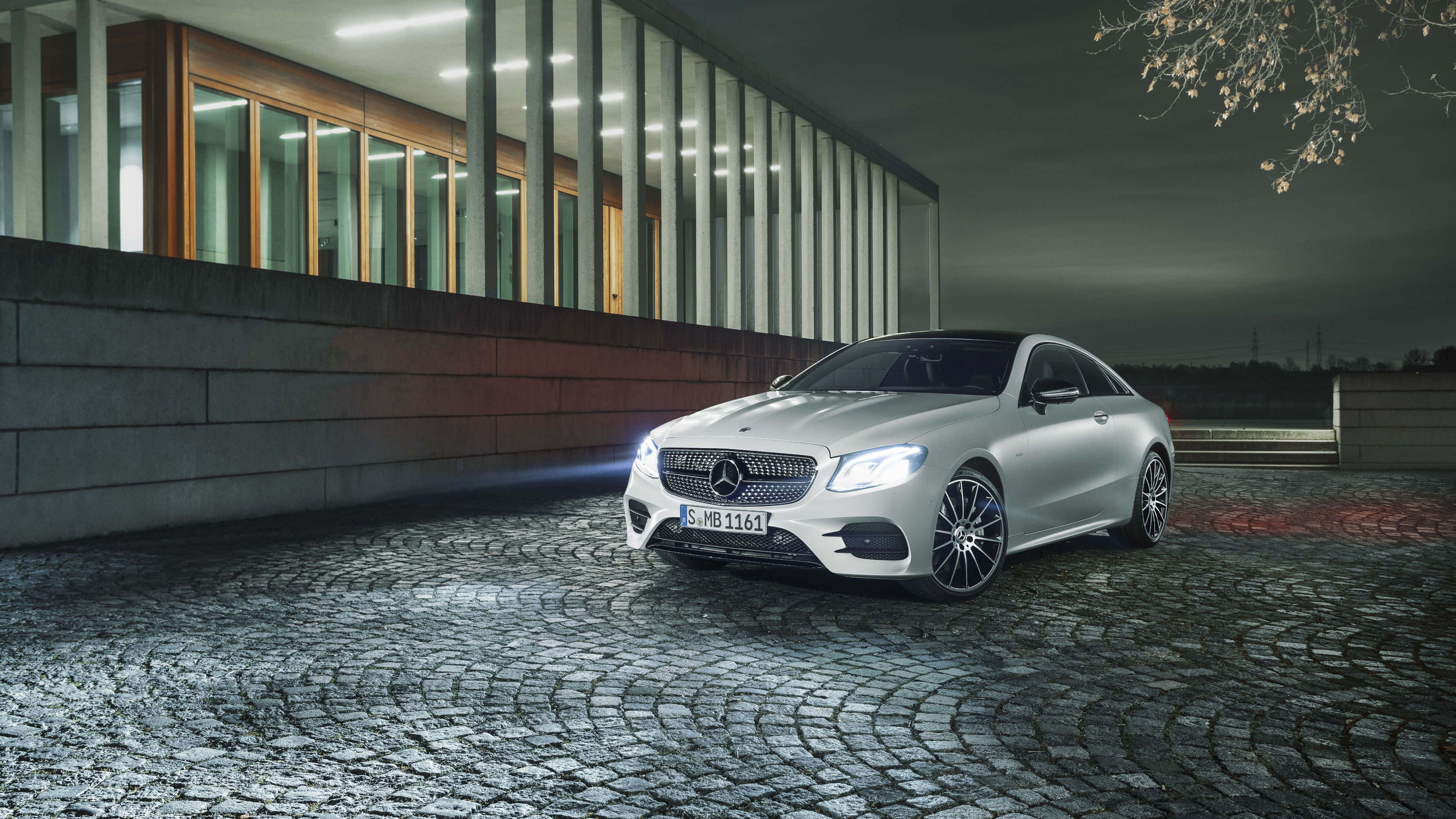 銀色梅賽德斯-奔馳 E 級 壁紙 — 一輛時尚的銀色梅賽德斯E級轎跑車在夜燈下，在現代玻璃建築旁的濕鵝卵石車道上閃閃發光。