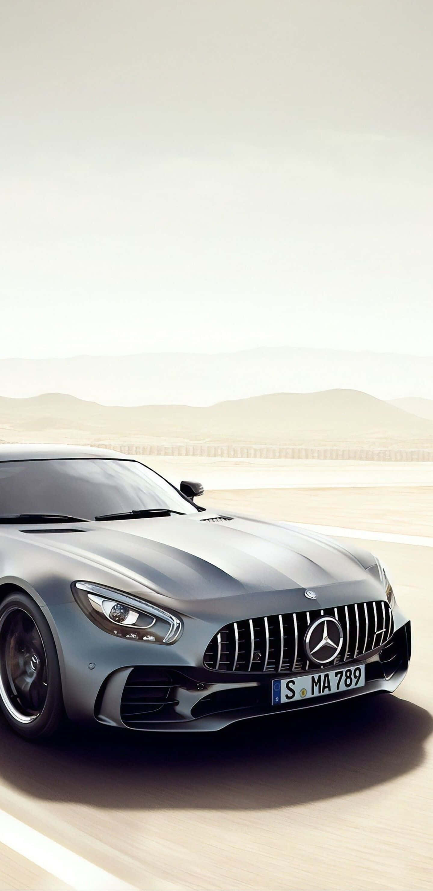 銀色梅賽德斯駛過沙漠道路 Pixel 3XL AMG GT-R 背景 壁紙 — 一輛流線型銀色 Mercedes-AMG GT 在沙漠公路上疾馳，遠山在朦朧的天空下若隱若現。
