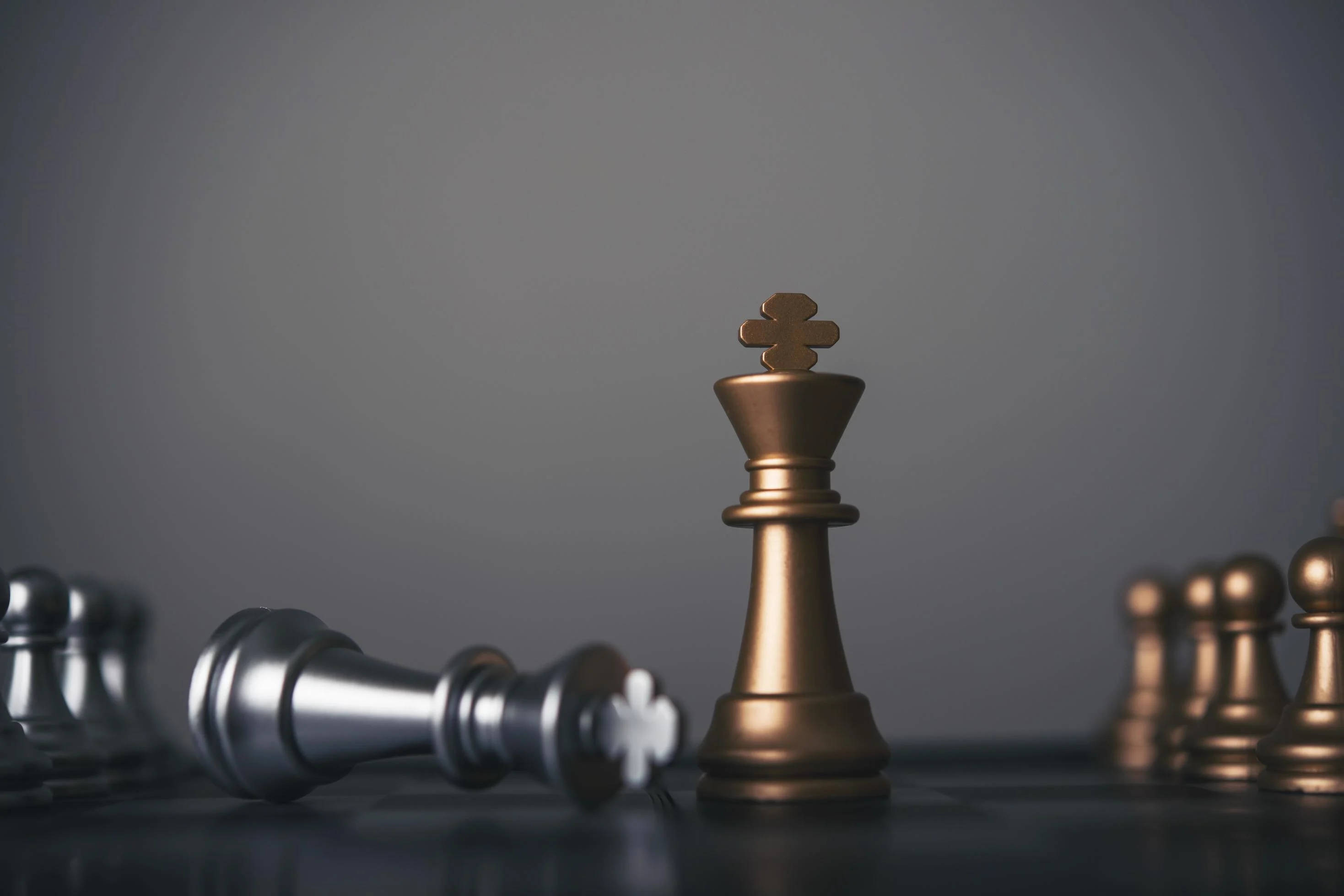 銀色棋王 壁紙 — 一隻金色嘅國際象棋國王高高企喺深色棋盤上，喺柔和灰色背景前，喺倒下嘅銀色棋子之上，充滿勝利姿態。