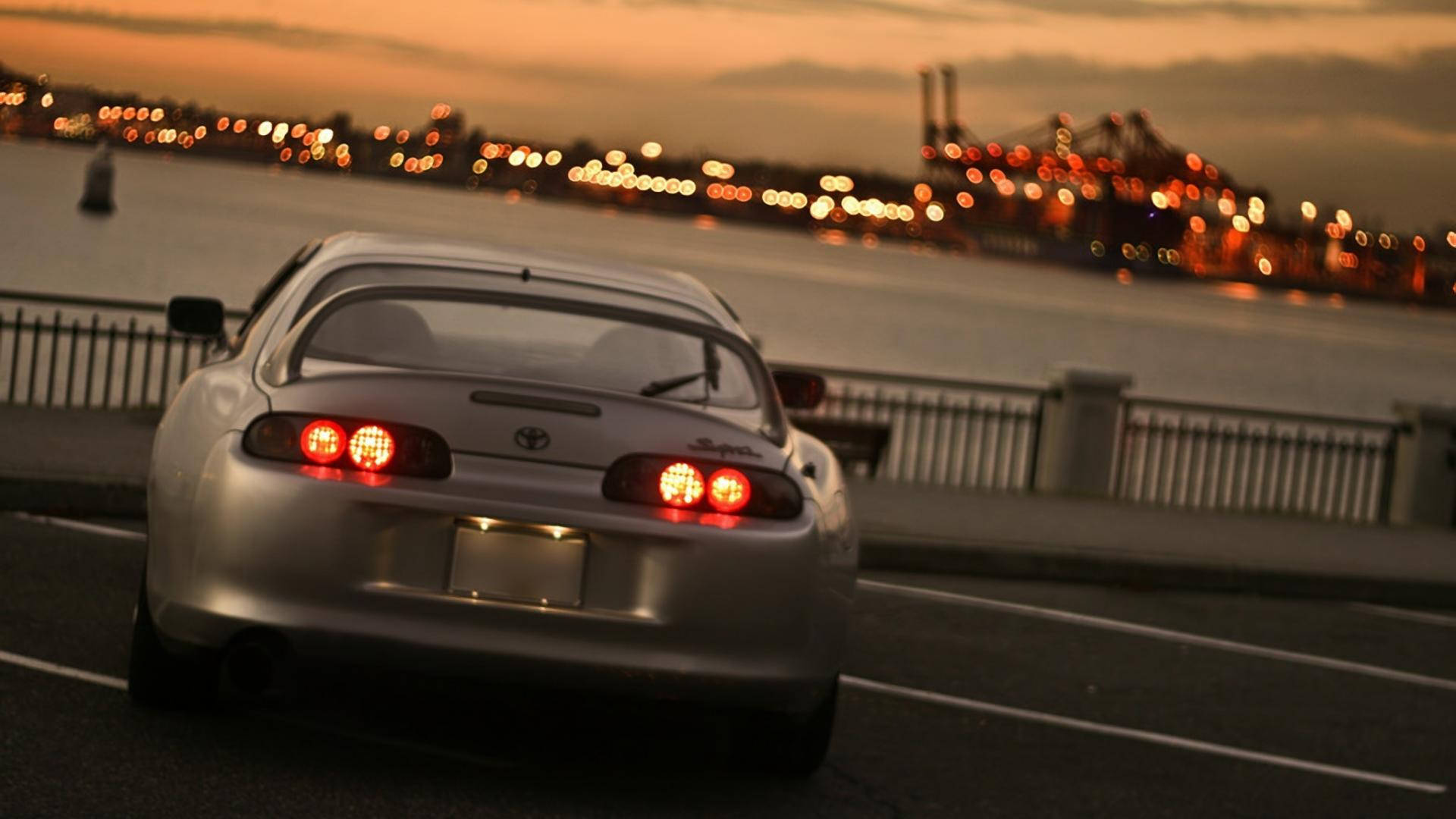 銀色跑車在黃昏時分在海濱附近行駛，背景城市燈光模糊。