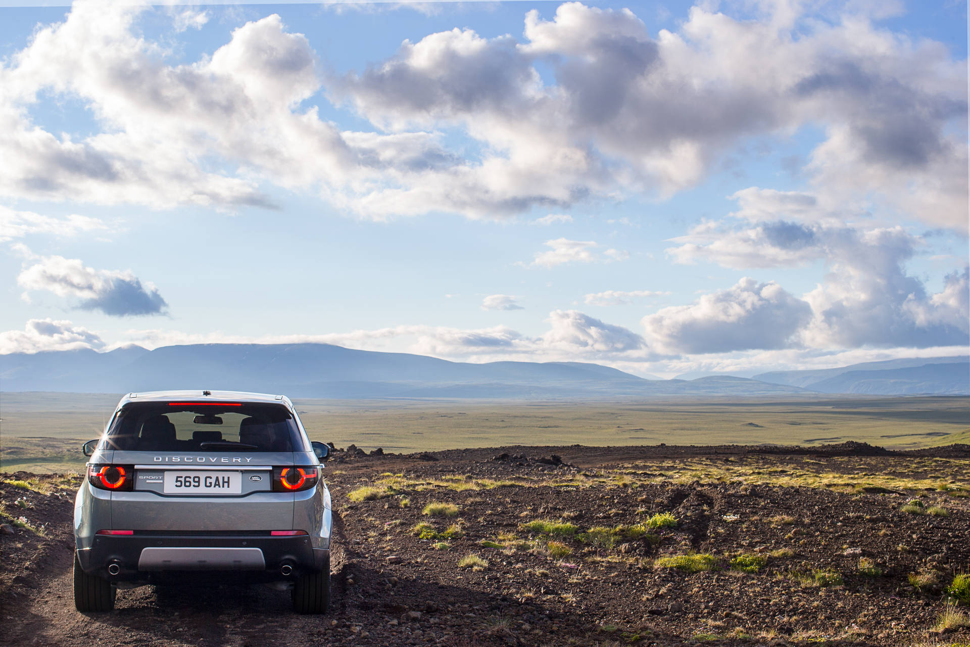 銀色 Land Rover Discovery 停在泥路上，面向開闊的風景，有山和雲。
