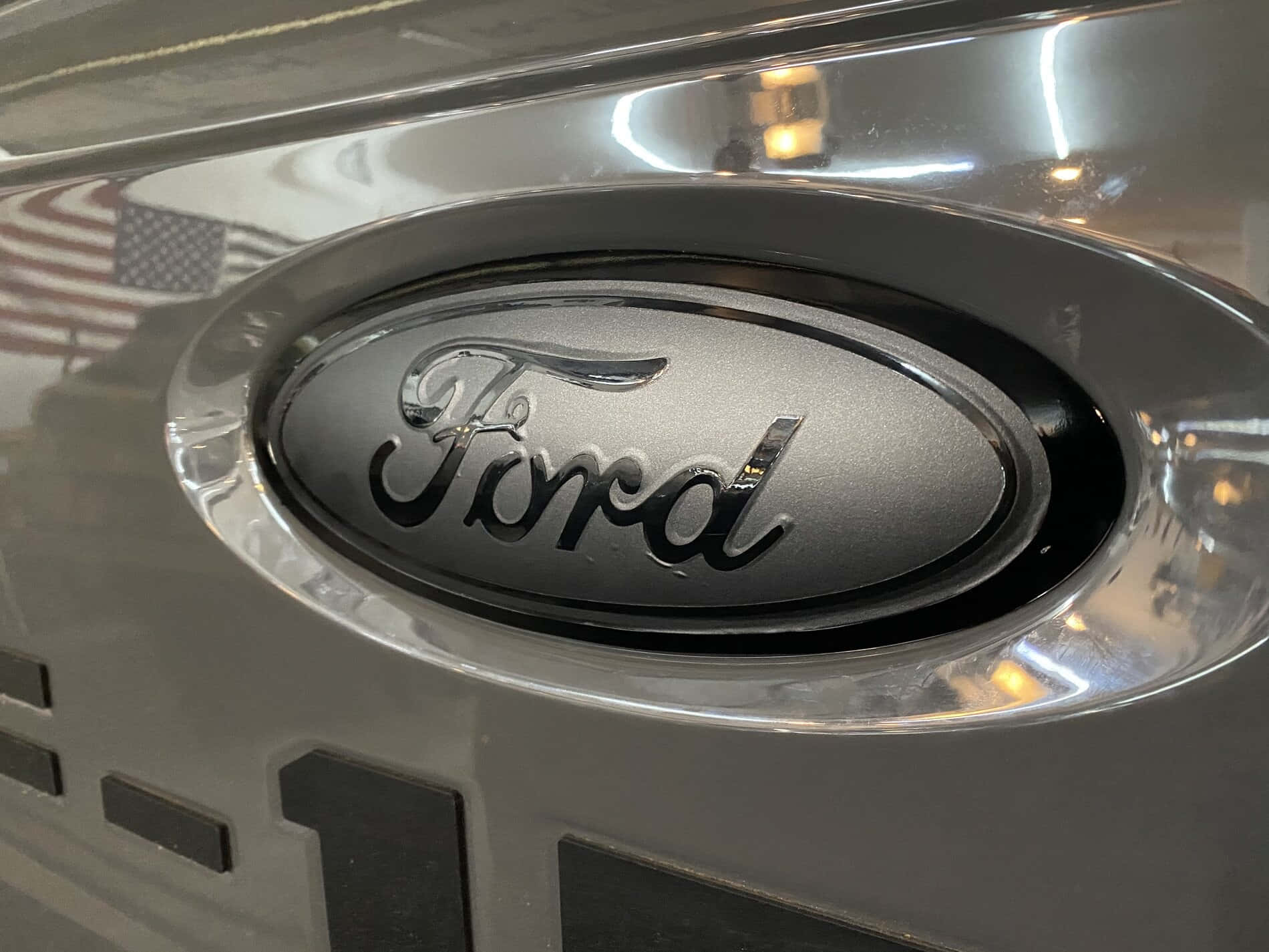Ford Logo 桌布 壁紙 — 銀色車頭蓋上鍍鉻福特標誌的特寫，反射美國國旗和室內燈光。