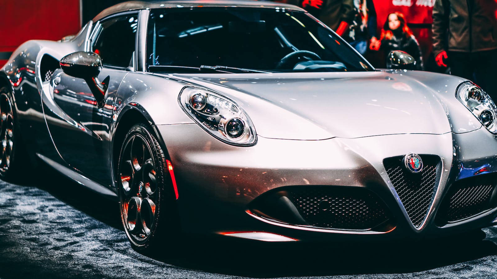 銀色阿爾法·羅密歐豪華跑車 壁紙 — 一輛時尚的銀色愛快羅密歐跑車在車展的明亮燈光下閃閃發光，背景是模糊的觀眾。