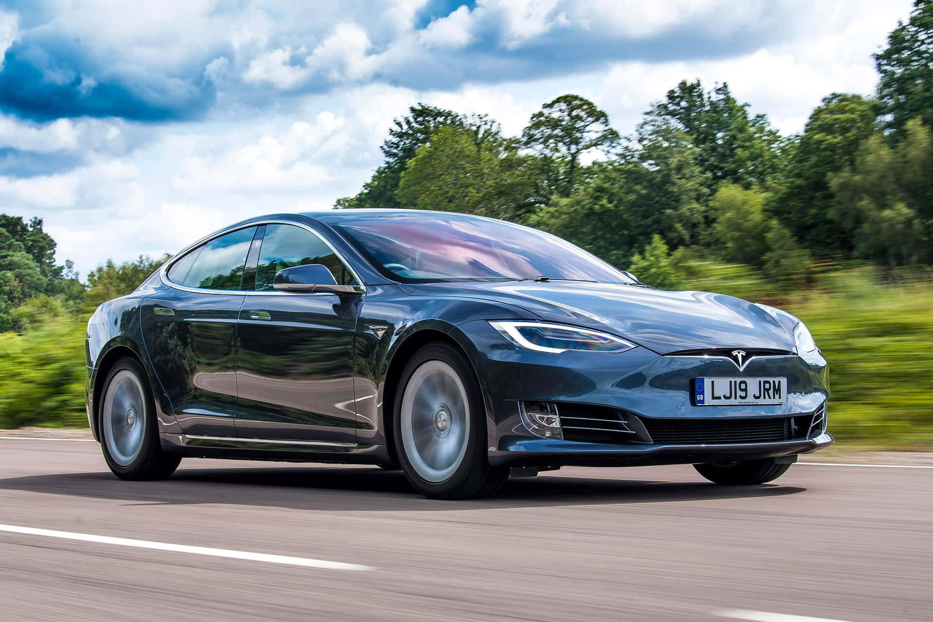 深藍色Tesla Model S在道路上奔馳，背景有綠色樹木和多雲天空。