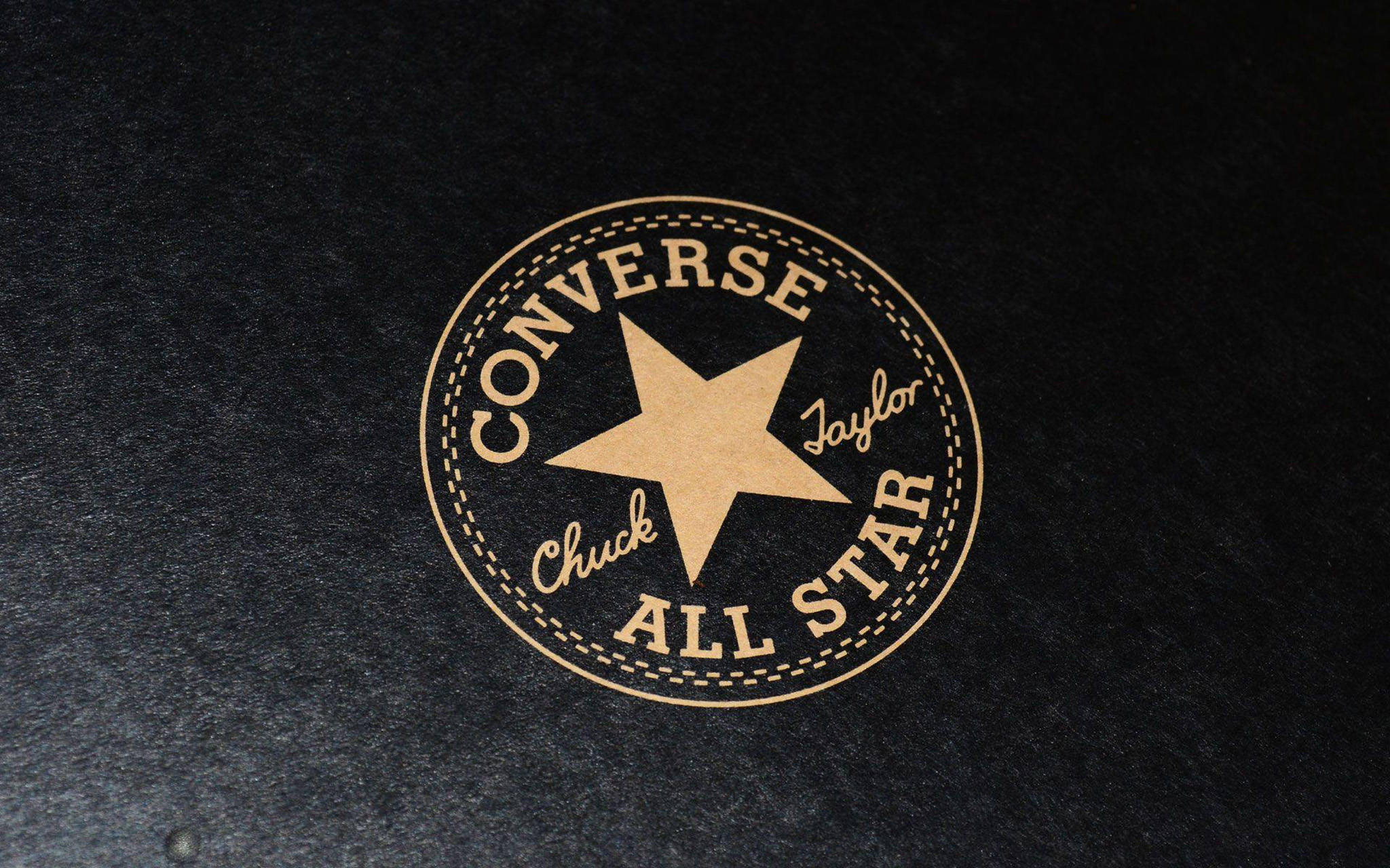 銅色 Converse 標誌 壁紙 — 一個復古的 Converse Chuck Taylor All Star 標誌，以棕褐色呈現於帶有紋理的黑色布料上，並附有縫線邊緣細節。