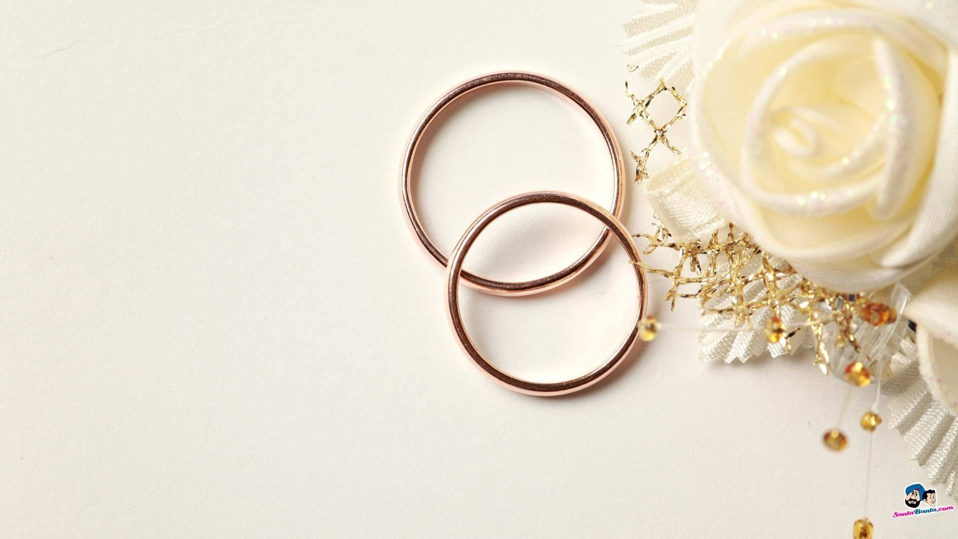 銅色婚戒 壁紙 — 兩隻玫瑰金結婚戒指放在白色玫瑰和金飾旁邊，置於潔白表面上。