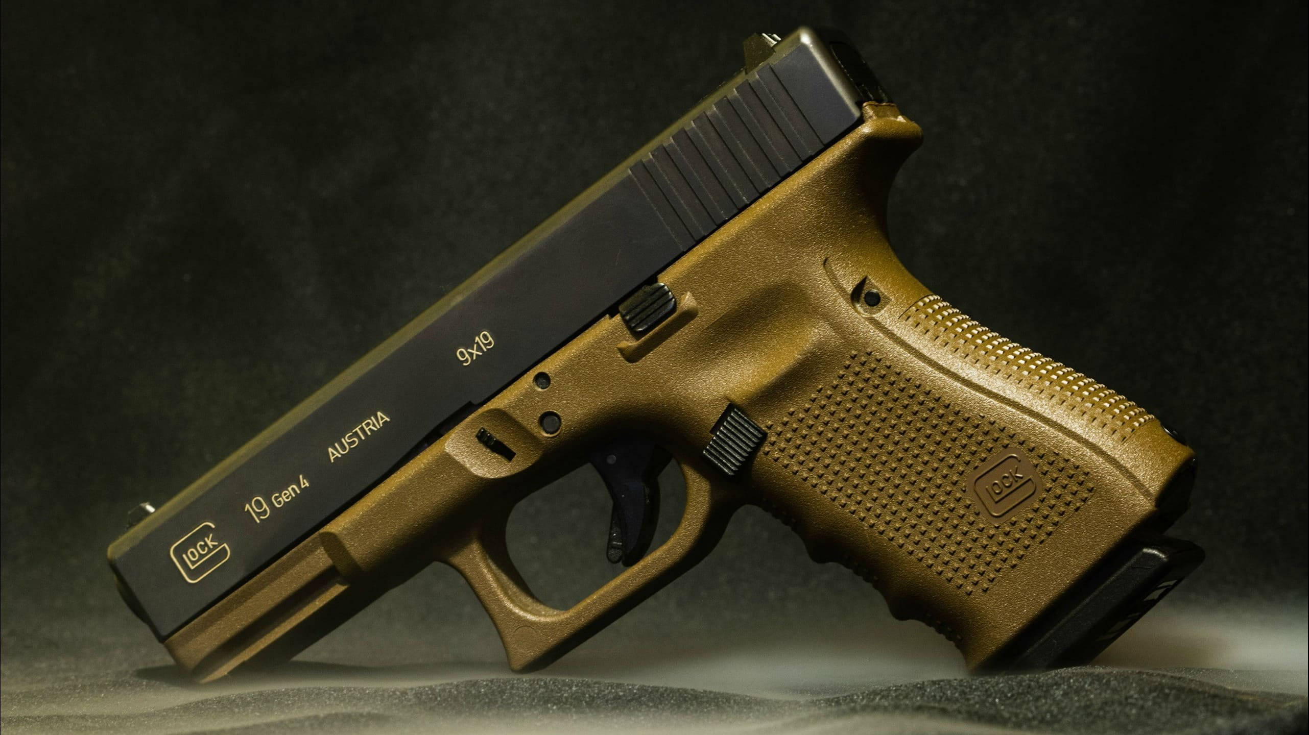 銅色格洛克19 Gen 4 壁紙 — 一把金色Glock 19 Gen4手槍對角放置在深色紋理表面上，在戲劇性的工作室燈光下。