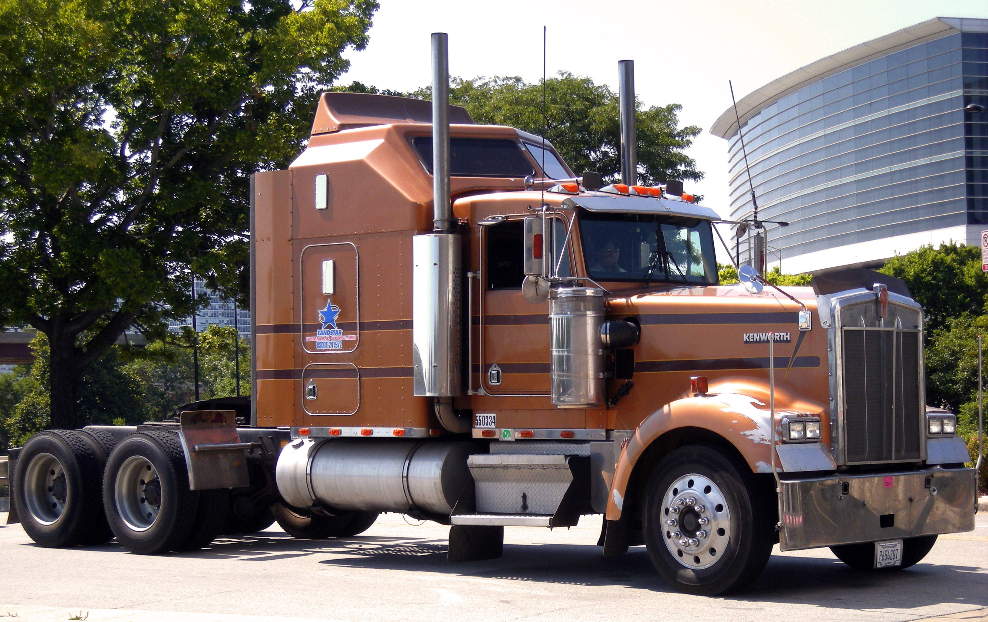 銅色肯沃斯 W900 壁紙 — 一輛帶有鍍鉻裝飾的大型橙色Kenworth半掛卡車停在城市街道上，旁邊有綠樹和現代玻璃建築。