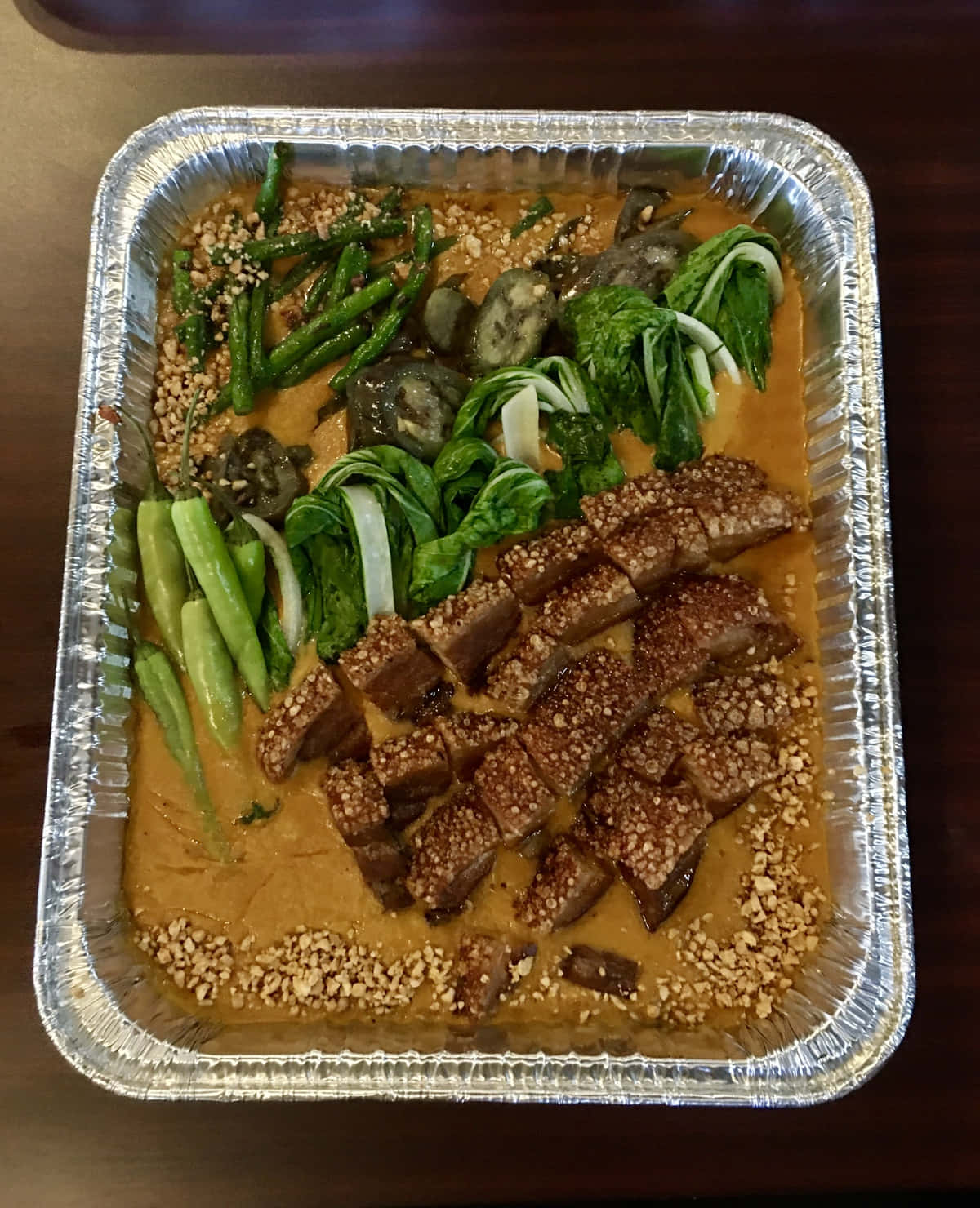 鋁箔盤盛裝的菲律賓燉菜 Kare-Kare 壁紙 — 一個錫紙托盤盛載著豐富的花生醬菜餚，配以脆皮五花腩、四季豆和白菜，並撒上芝麻作裝飾。