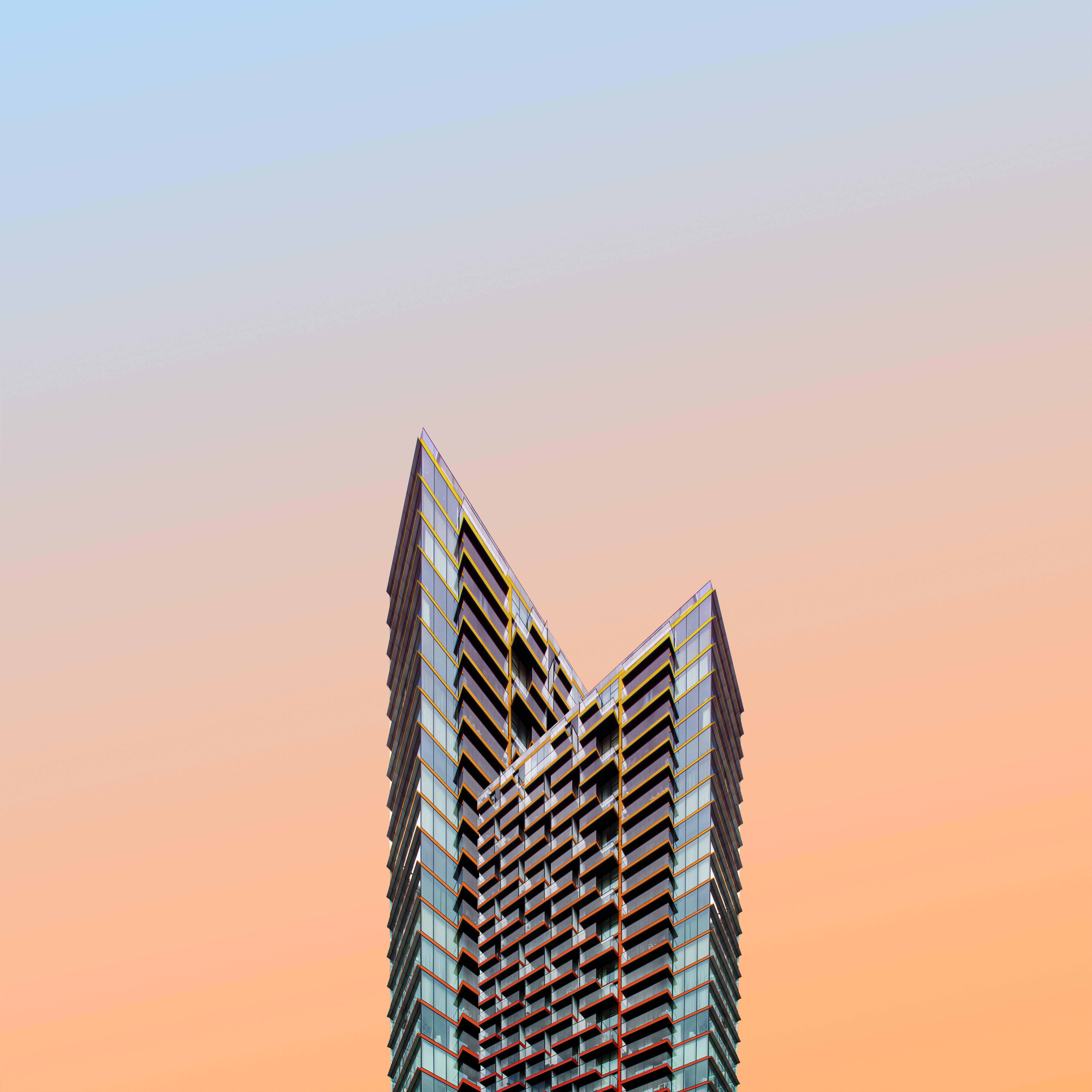 鋒利邊緣高樓建築畫作 壁紙 — 一座現代玻璃摩天大樓，帶有幾何陽台，刺穿從柔和藍色過渡到溫暖桃色的漸變天空。