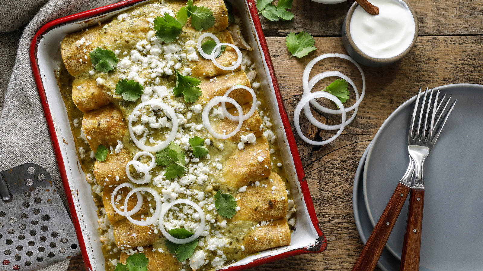 鋪有洋蔥圈的 enchiladas 盤 壁紙 — 金黃色的玉米捲餅浸在綠色醬汁中，上面撒有洋蔥、香菜和碎奶酪，盛在紅色琺瑯盤中，放在質樸的木桌上。