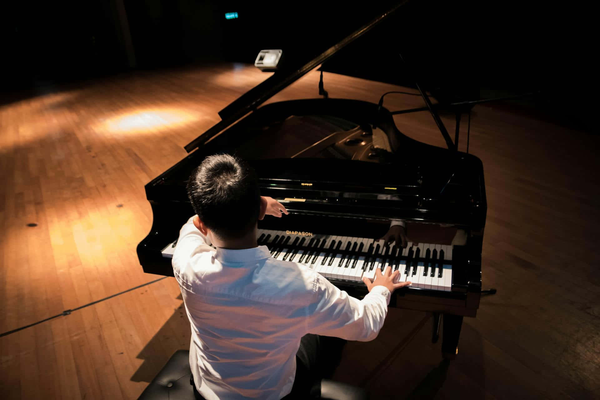 鋼琴家於音樂會舞台演奏 壁紙 — 一位身穿白襯衫的鋼琴家在昏暗的音樂廳木質舞台上，在溫暖的聚光燈下彈奏三角鋼琴。