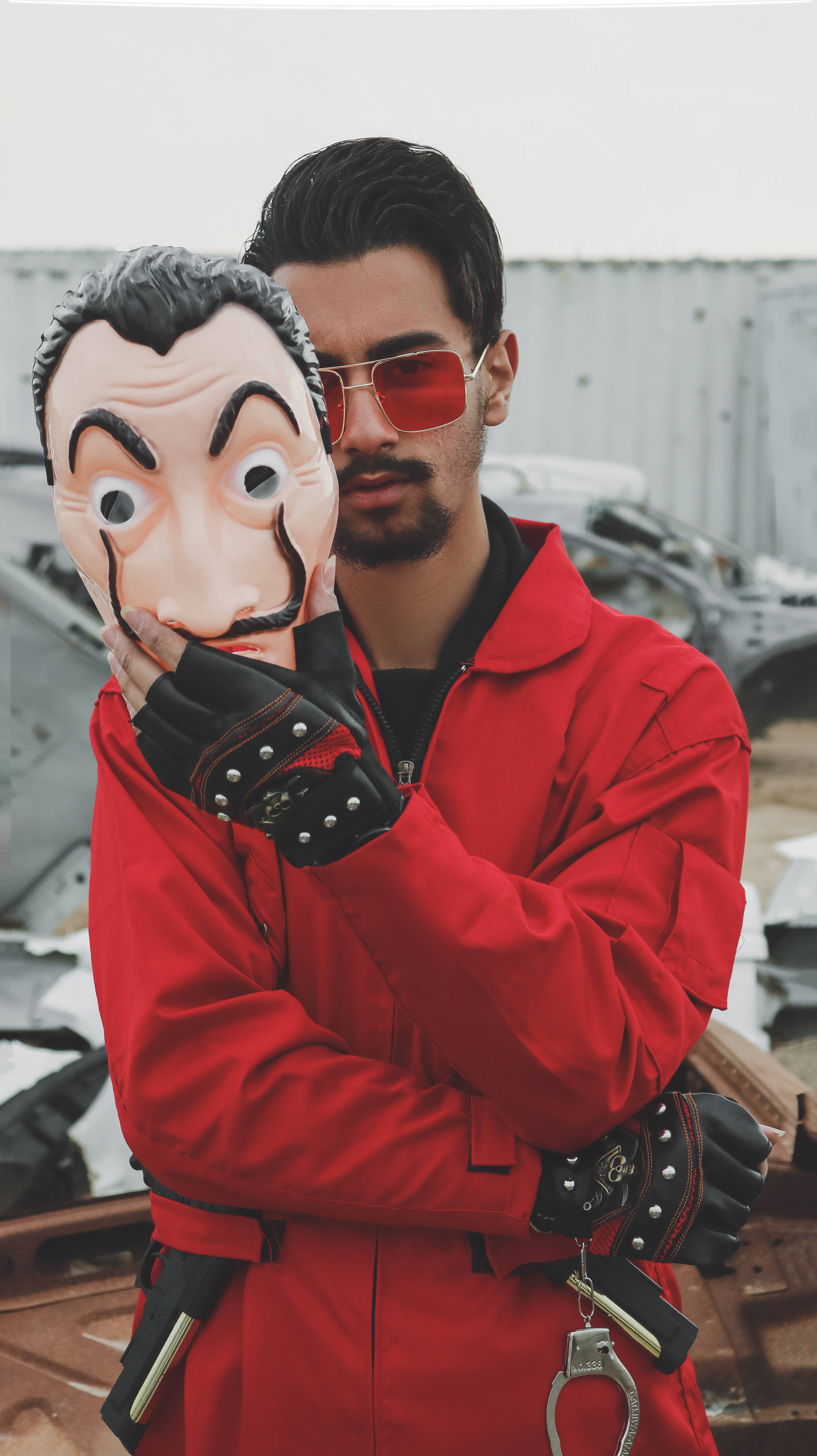 《錢他》面具cosplay 壁紙 — 一個身穿紅色連身衣和紅色太陽眼鏡的男人，站在廢車場，手持薩爾瓦多·達利面具。