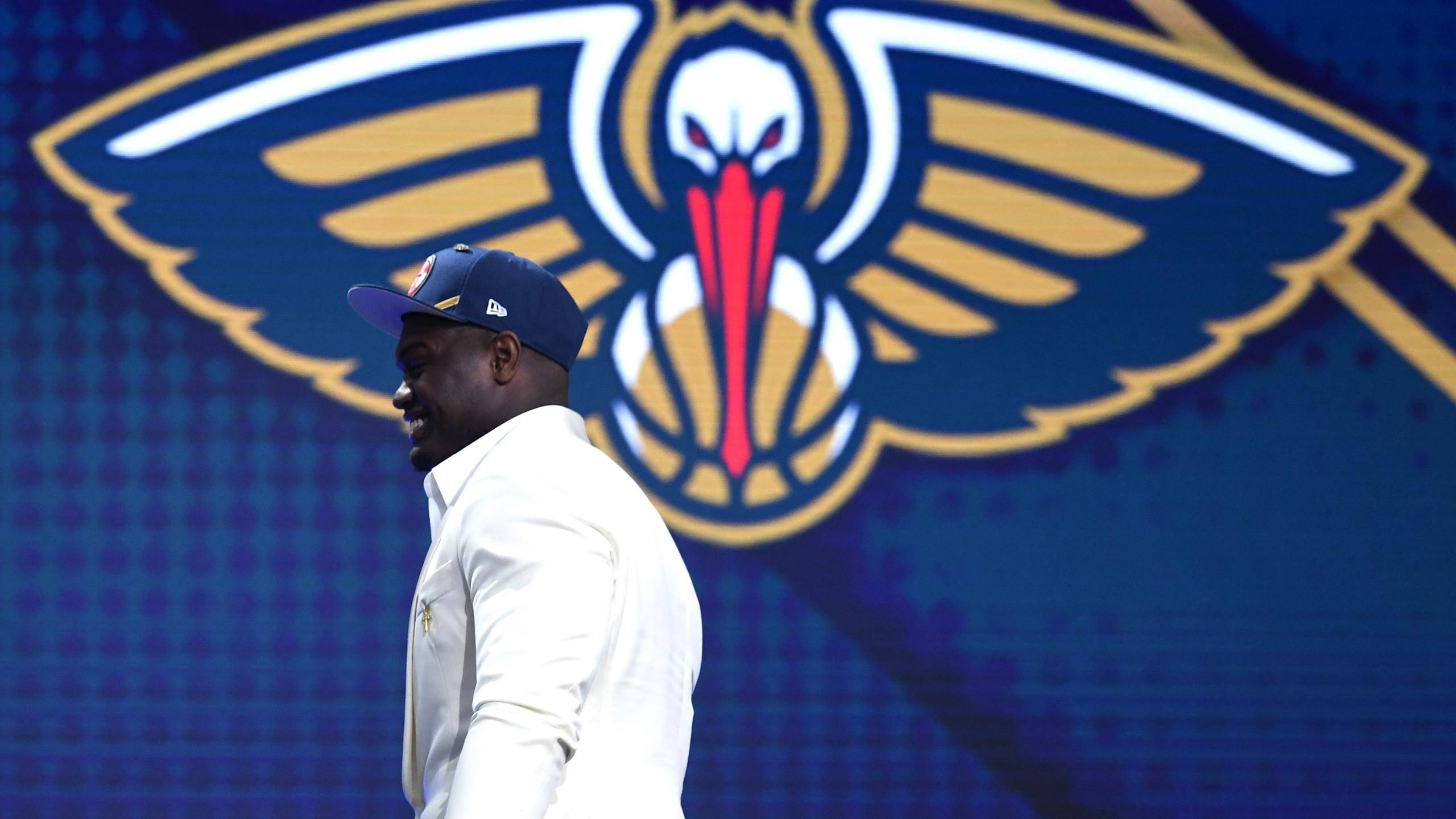 錫安·威廉森 2019 NBA選秀 壁紙 — 一名身穿白色西裝和藍色帽子的男子走過一個藍色數碼屏幕上巨大的新奧爾良鵜鶘籃球隊標誌。
